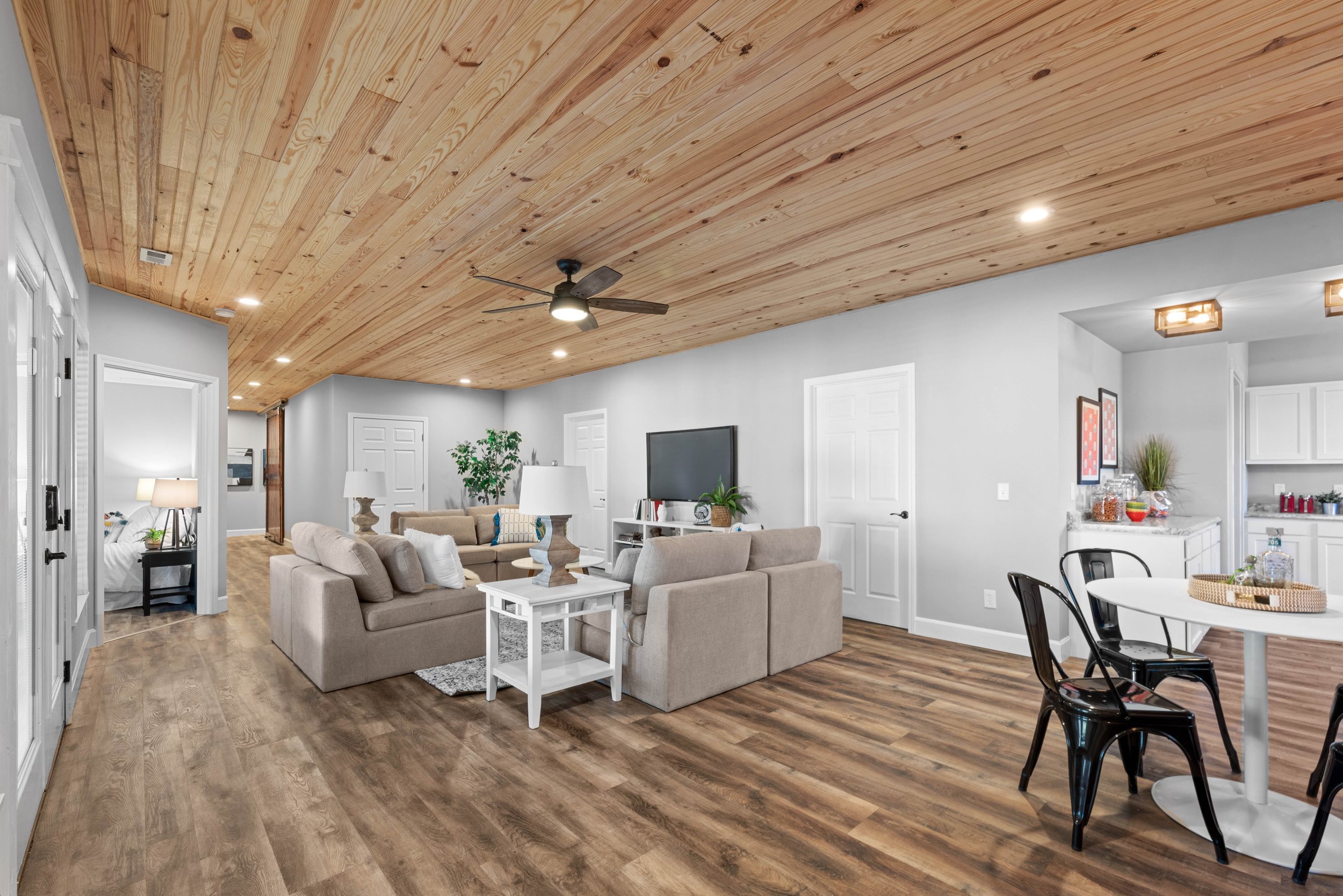 Spacious living room with wooden ceiling, beige sectional sofa, flat-screen TV, wooden flooring, and a small dining table with black chairs.