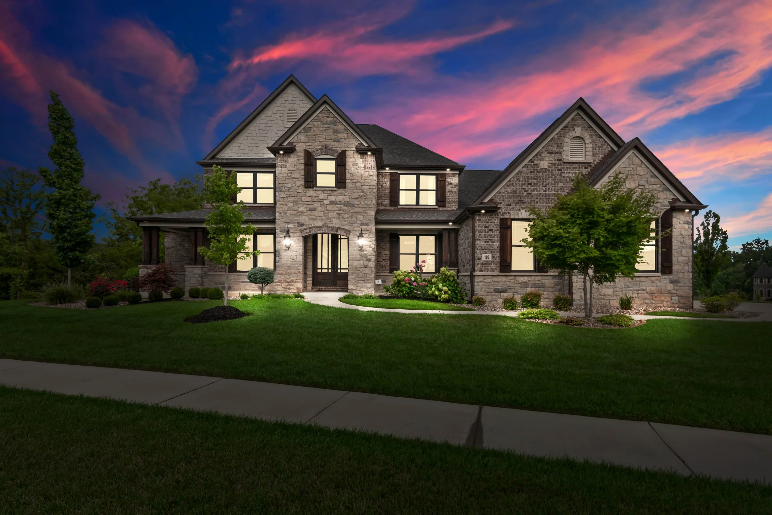 A large brick and stone house with multiple gabled roofs, illuminated in the evening, with a well-manicured lawn and trees in front, under a colorful sunset sky.