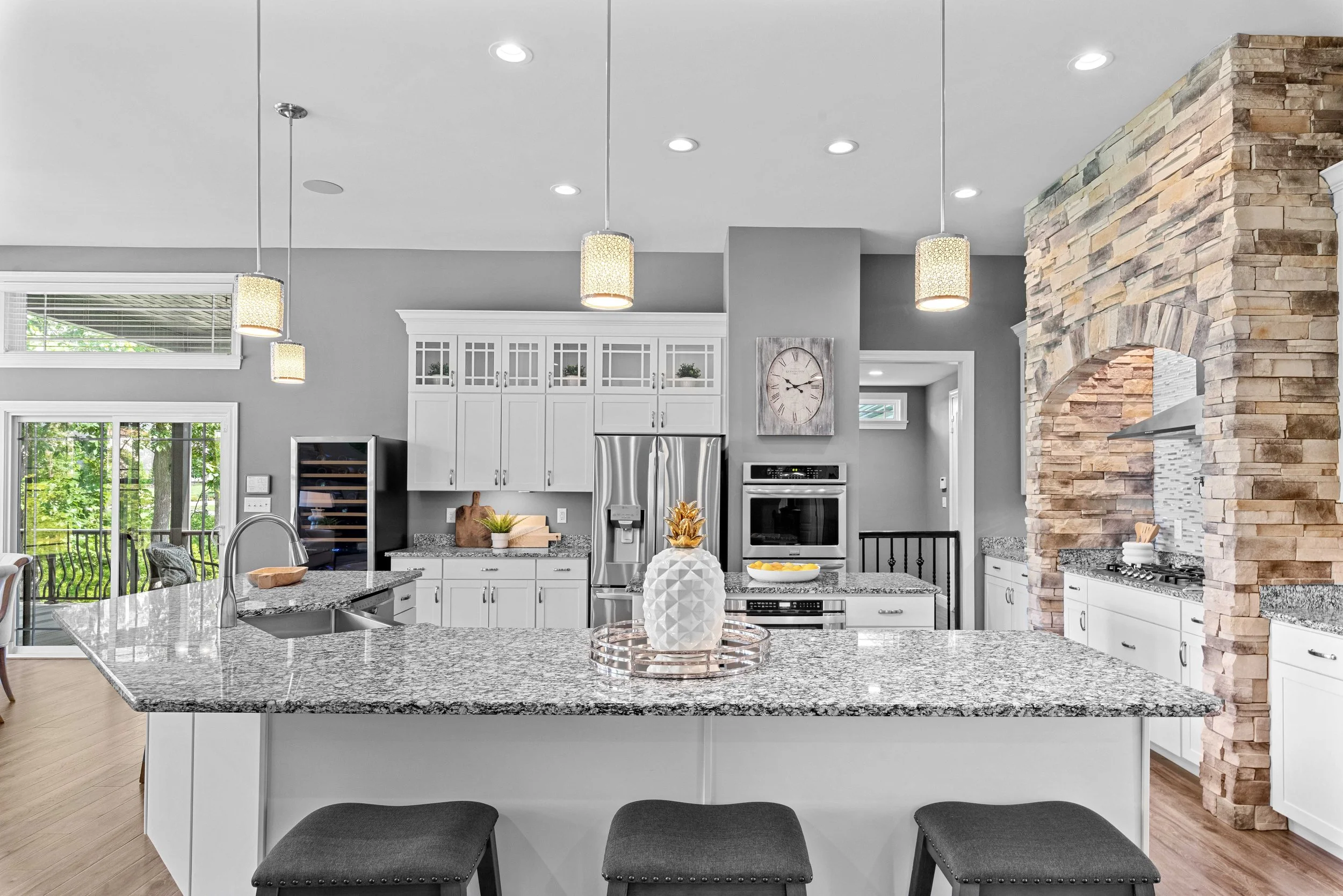 Modern kitchen with white cabinets, granite countertops, stainless steel appliances, and a stone accent wall. There is a island with bar stools, decorative pineapple, and dining area with sliding glass doors showing outdoor greenery.