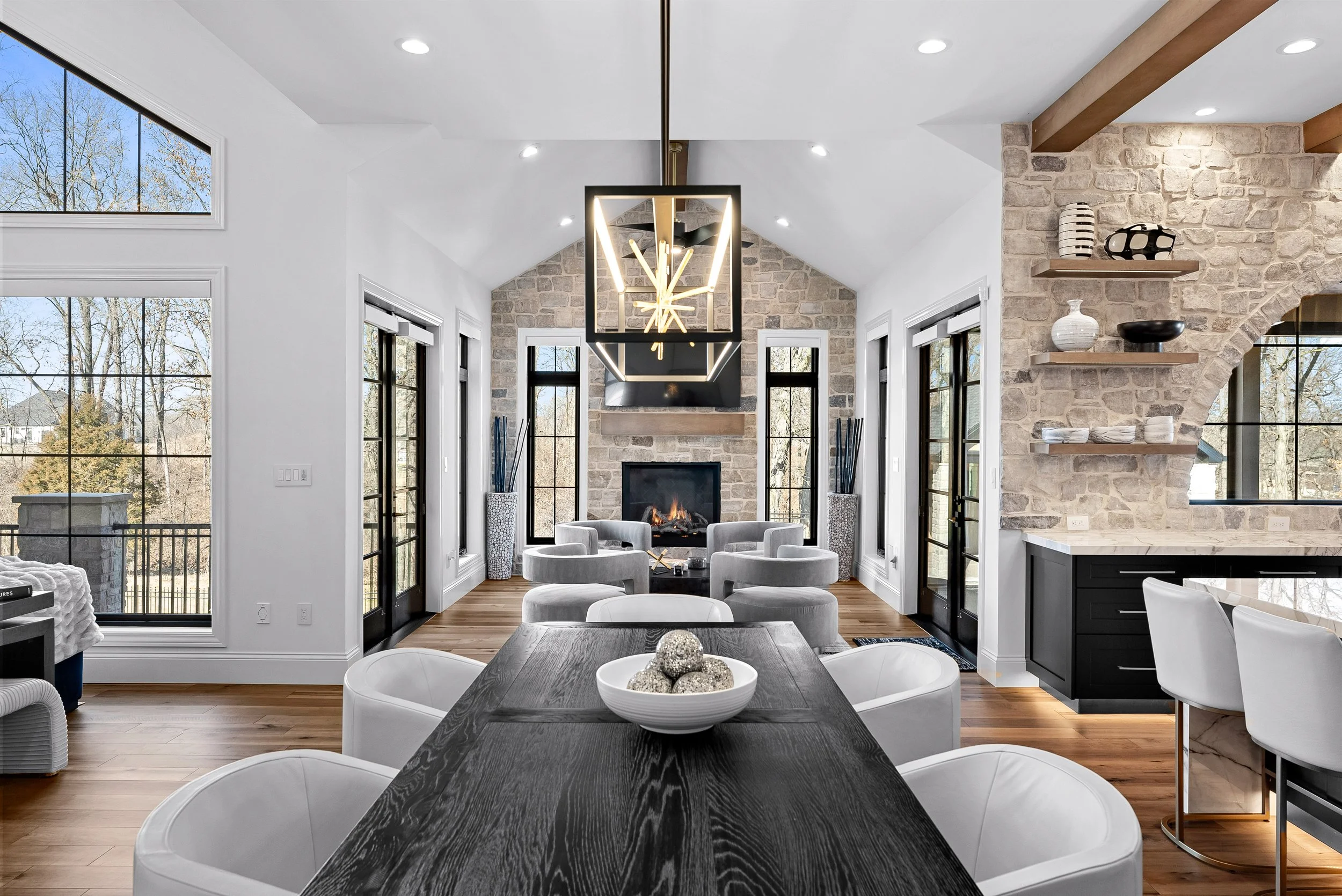 Modern living room with a fireplace, gray chairs, and large windows showing trees outside. A wooden dining table with white chairs and a bowl of decorative balls, and a kitchen area with black cabinets and floating shelves with vases and bowls.