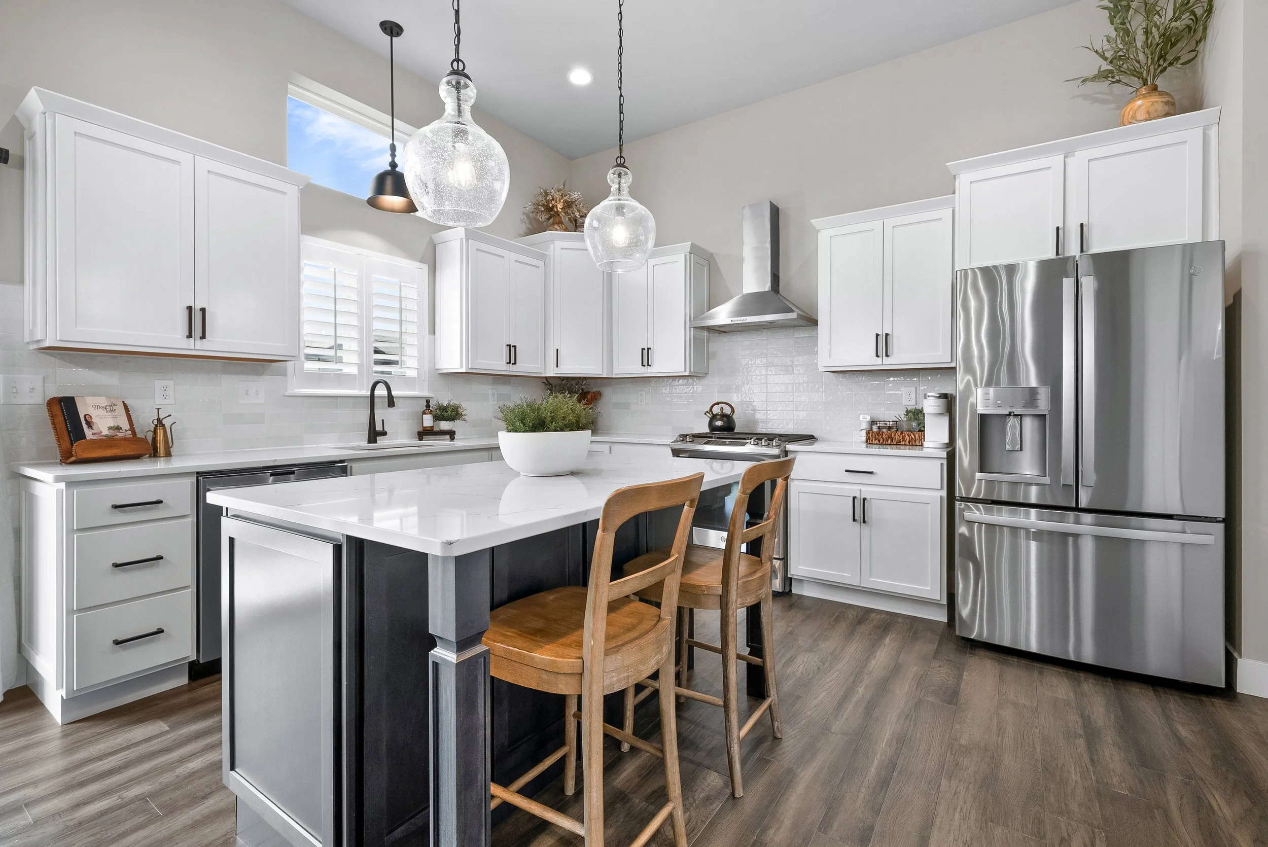 Modern farmhouse kitchen with white cabinets, stainless steel refrigerator, island with a dark base and white countertop, two wooden chairs, pendant lights, and a window with white shutters.