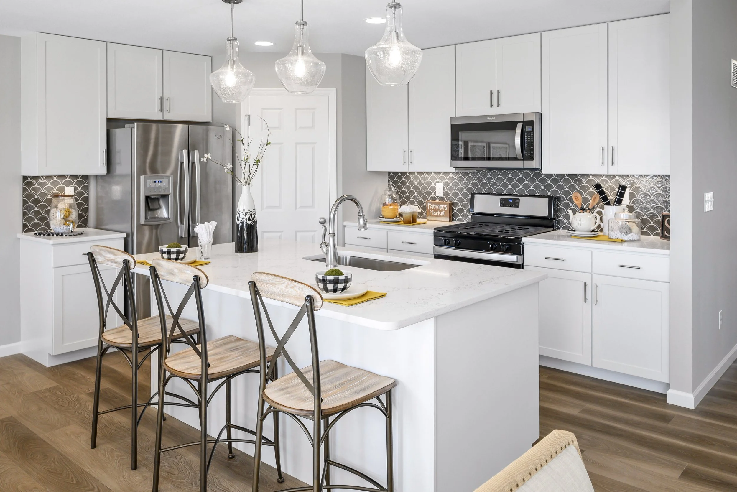 Modern kitchen with white cabinets, stainless steel appliances, a marble island with three chairs, pendant lights, grey backsplash, and wood flooring.