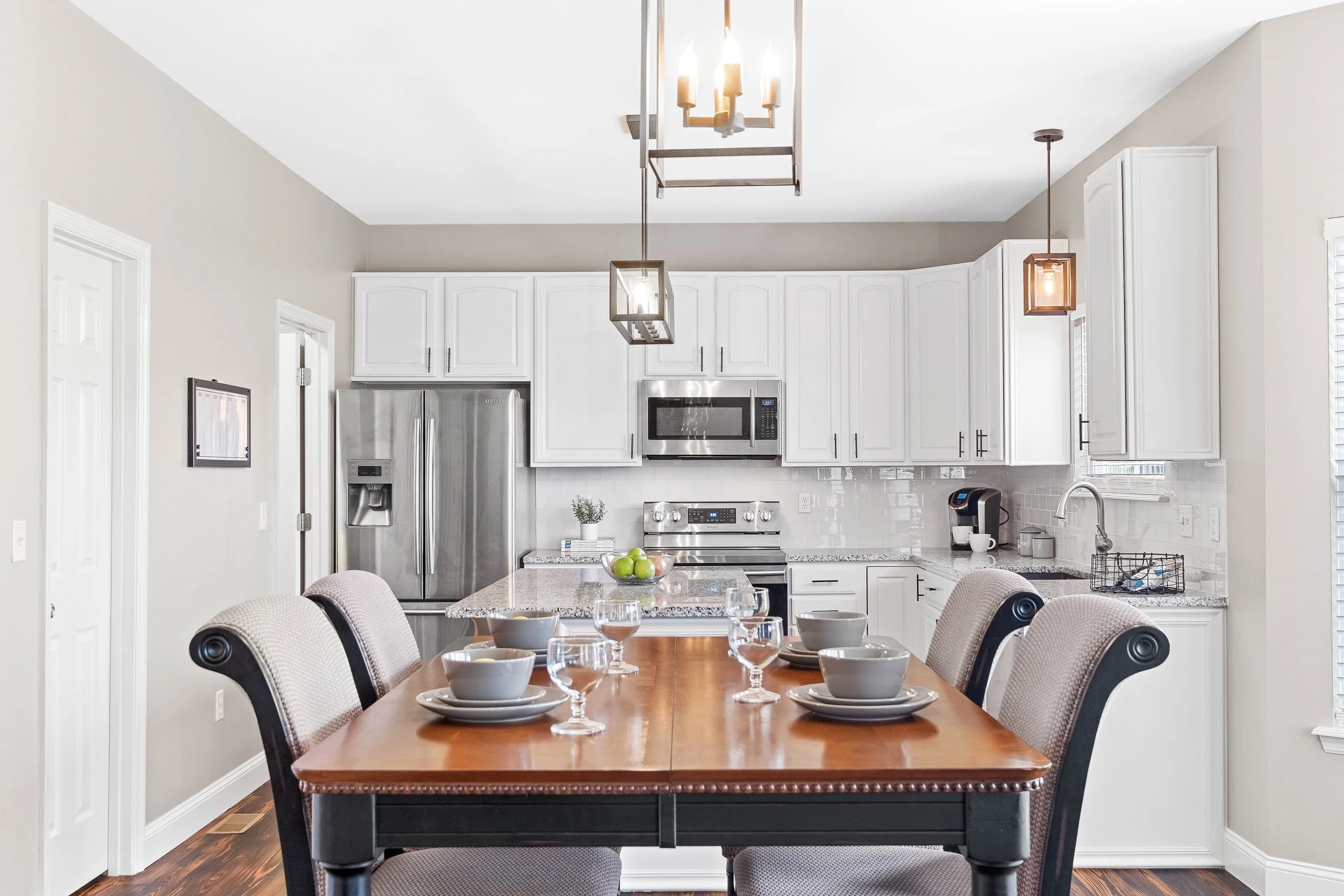 Modern kitchen with white cabinets, stainless steel appliances, granite countertops, dining table with place settings, and contemporary lighting fixtures.