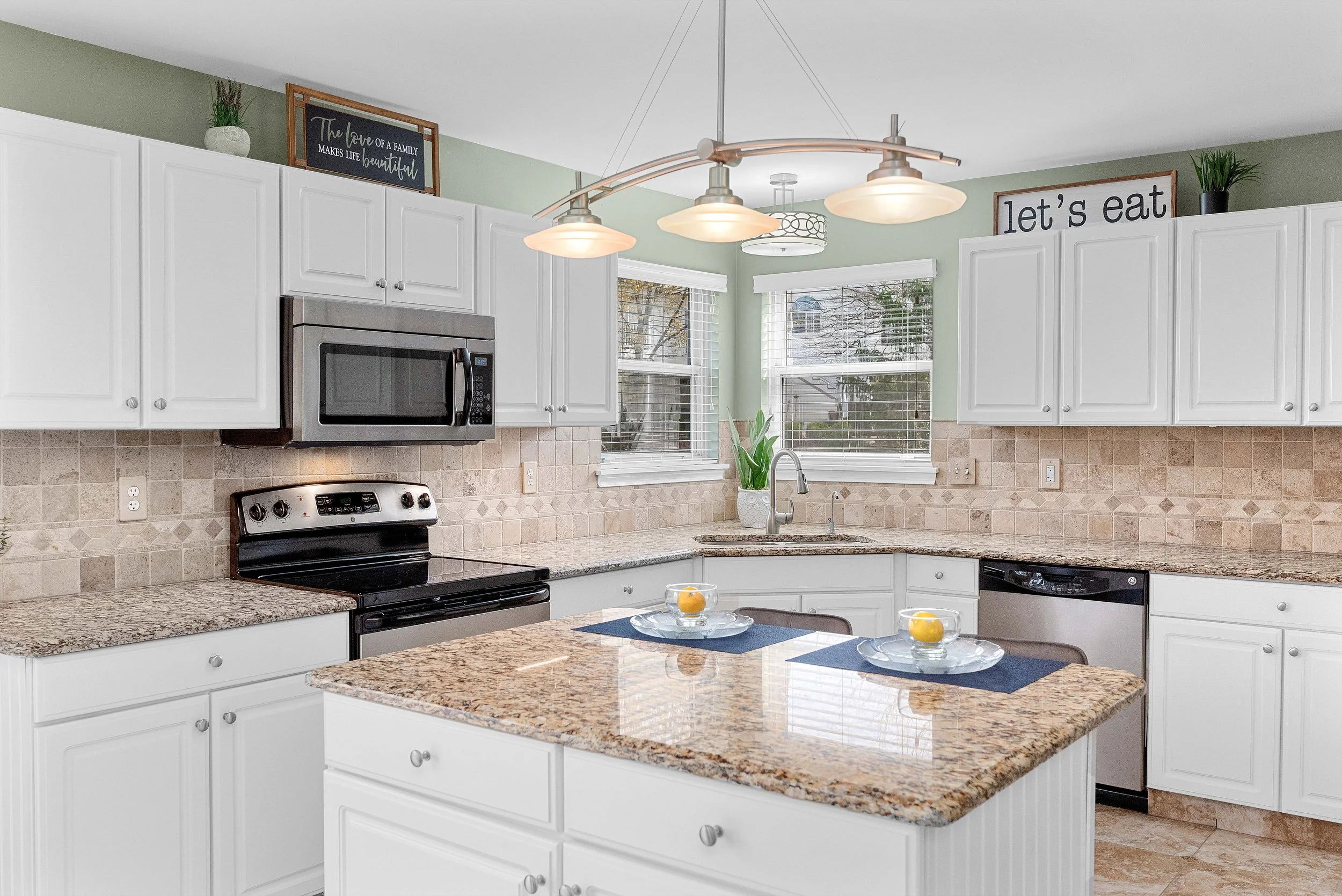 Bright kitchen with white cabinets, granite countertops, beige tile backsplash, and stainless steel appliances including microwave and oven. There are two large windows with white blinds, a kitchen island with placemats and glass bowls, and decorativ