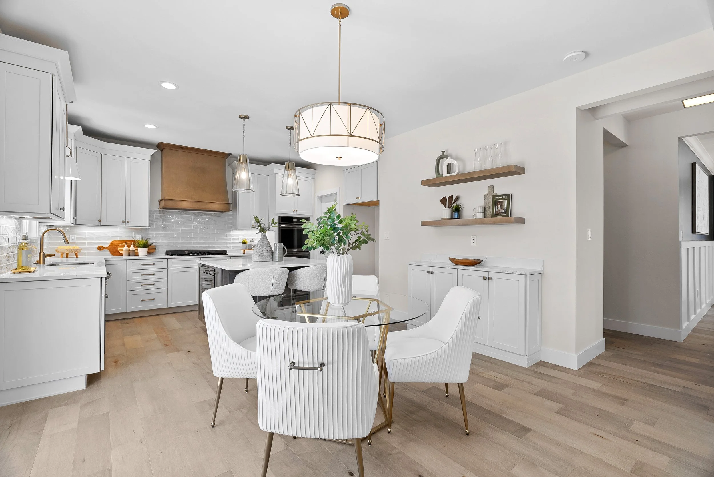 Modern kitchen and dining area with white cabinetry, gold accents, light wood flooring, and a round glass table with white chairs.