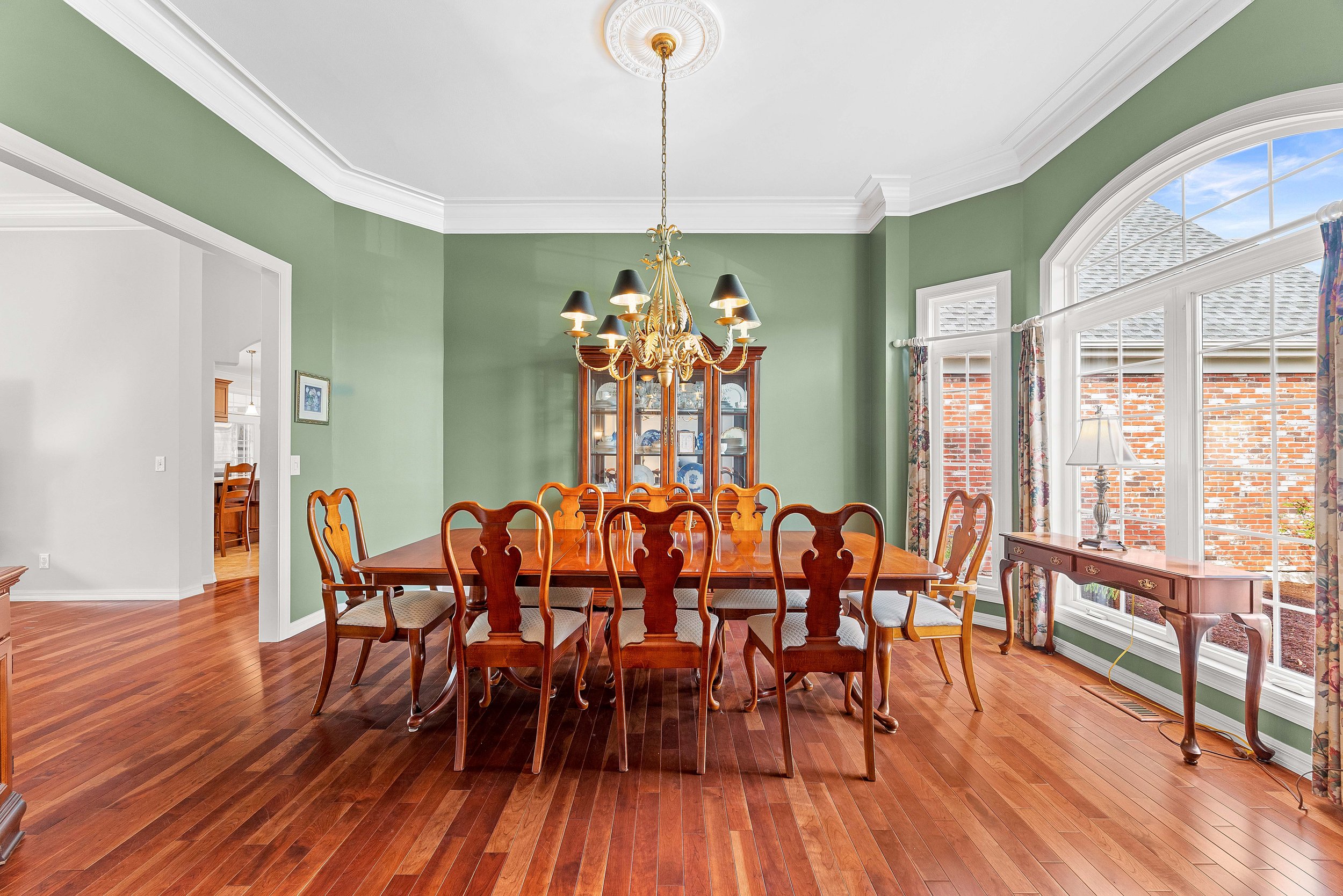 A dining room with green walls, wooden floor, and a large rectangular wooden dining table with chairs. There is a chandelier above the table and a china cabinet in the background. Large windows with floral curtains and a side table with a lamp.