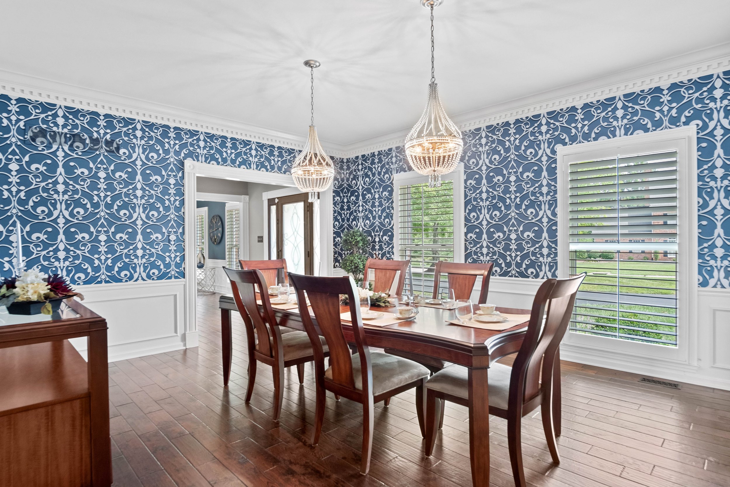 Elegant dining room with a wooden table, eight chairs, two hanging chandeliers, large windows with white shutters, blue and white patterned wallpaper, and hardwood floors.