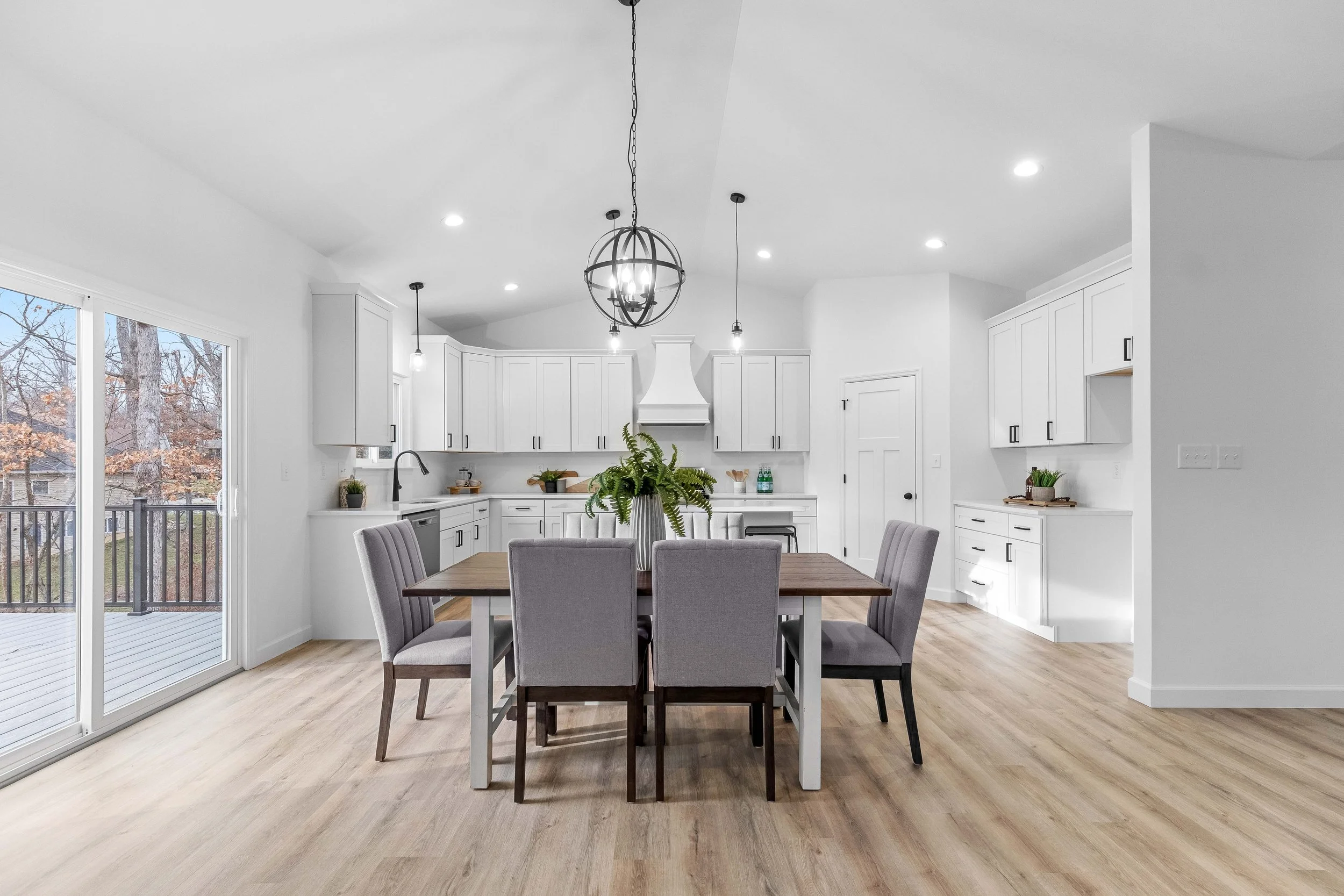 Modern open-concept kitchen and dining area with white cabinets, wood flooring, a sliding door to an outdoor deck, and a wooden dining table surrounded by six gray upholstered chairs.