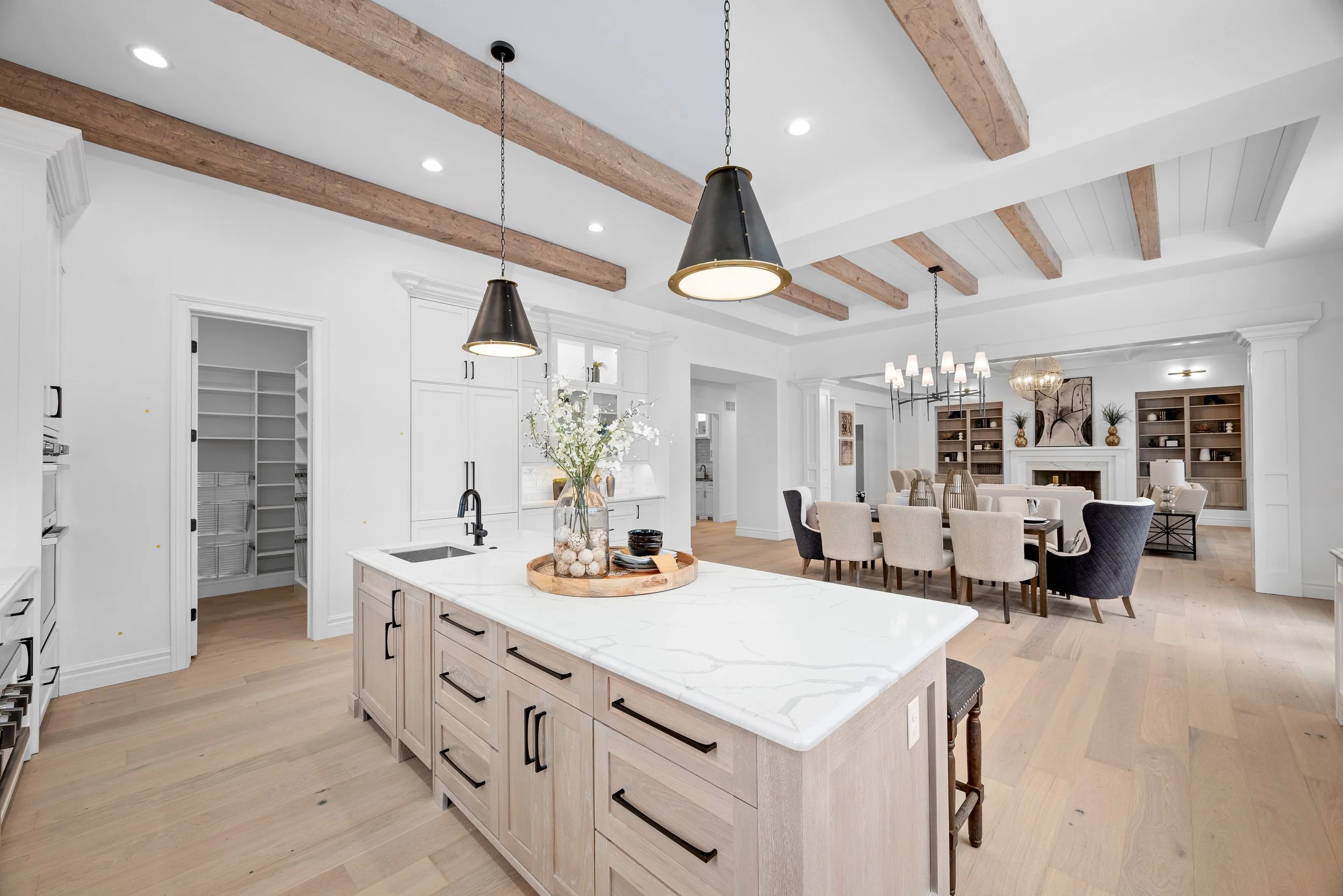 Open-concept kitchen and dining area with white walls, light wood flooring, a kitchen island with a marble top, black pendant lights, and a formal dining table with cream and dark blue chairs. There's a large chandelier and built-in bookshelves in th