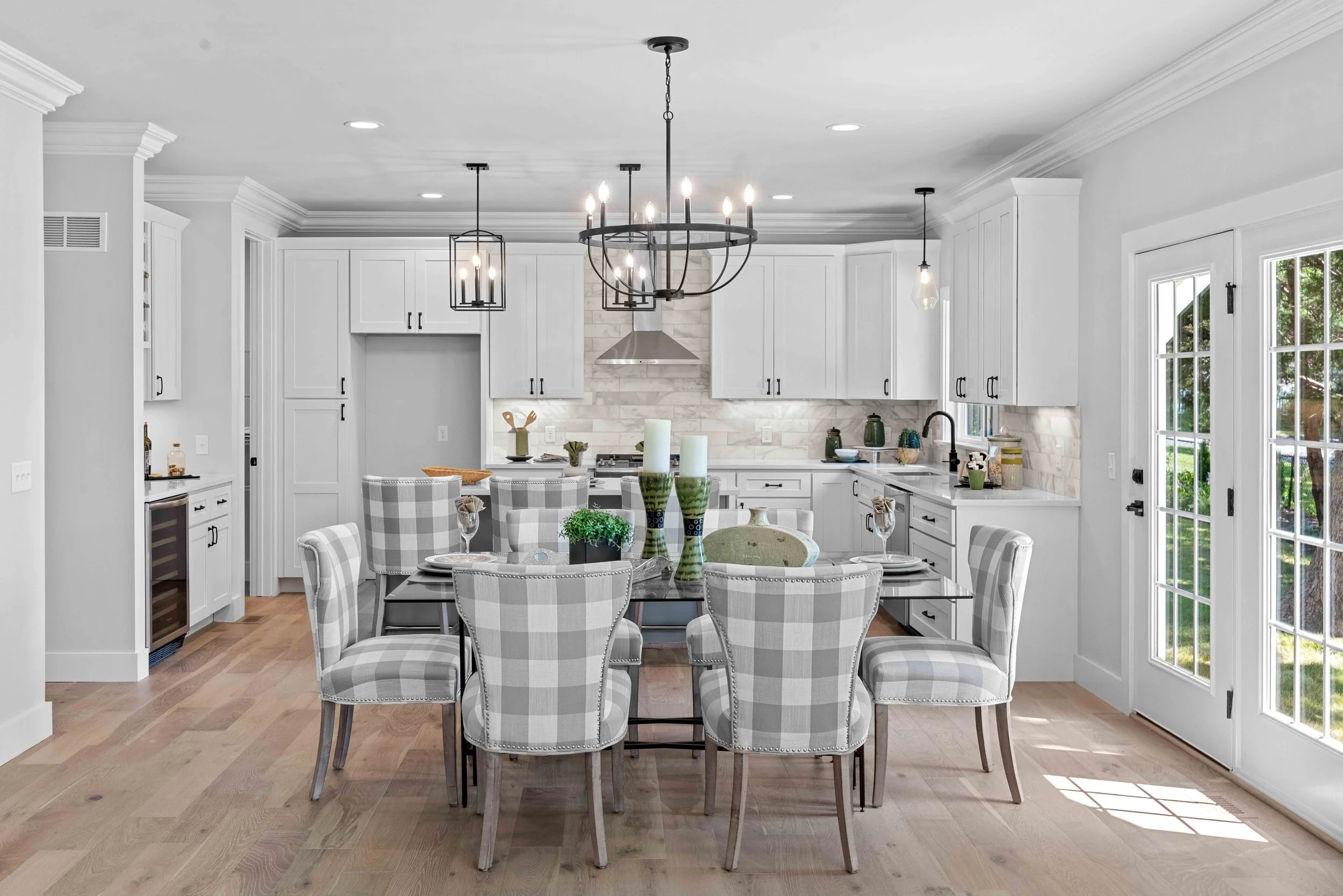Bright, white kitchen with a round dining table surrounded by checkered chairs, modern pendant and chandelier lighting, white cabinets, light hardwood floors, and large windows with a view of greenery outside.