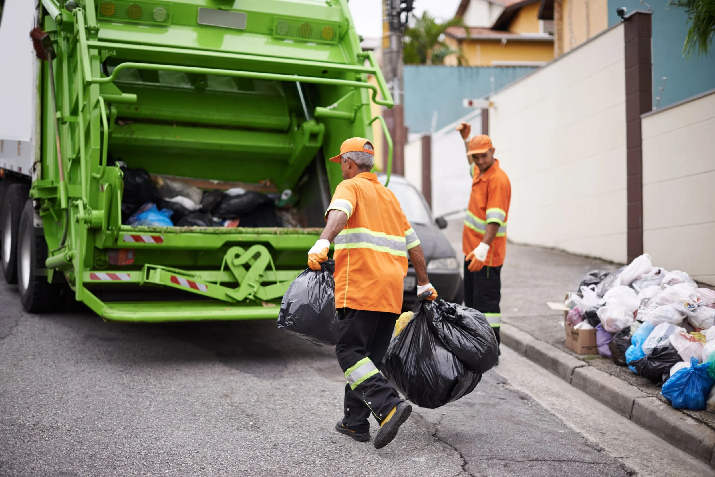 Two sanitation workers in orange uniforms and gloves collecting trash bags near a green garbage truck in an urban neighborhood, with trash on the ground