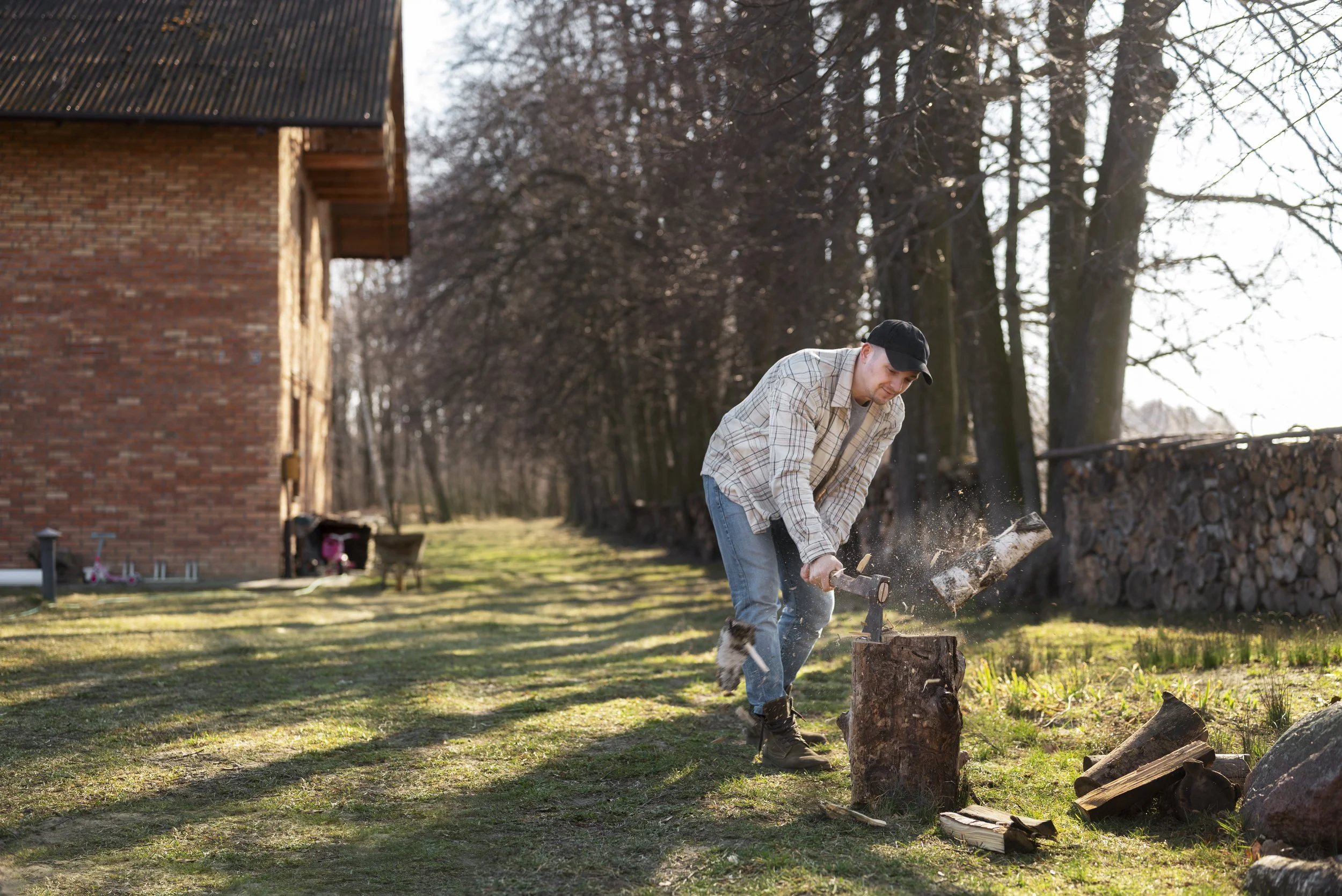 A man wearing a plaid shirt, blue jeans, and a black cap chopping wood outside near a brick house and a row of trees.