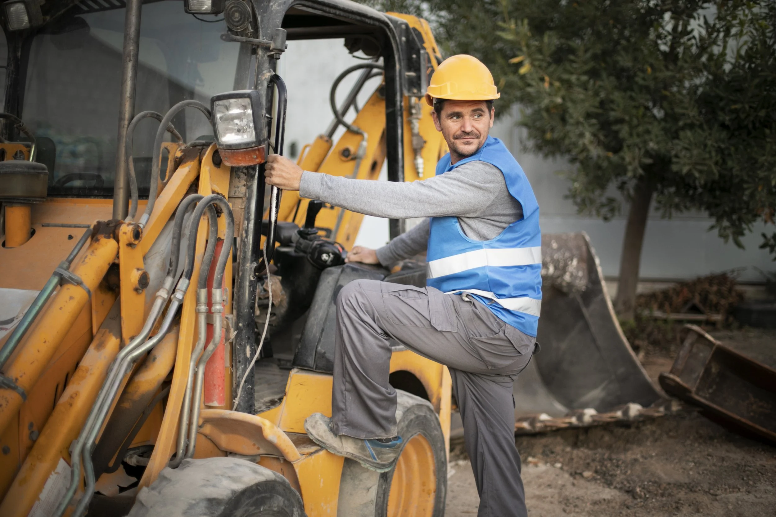 A male construction worker wearing a yellow safety helmet, blue safety vest, and gray pants, standing next to a yellow backhoe loader, with one foot on the machine's step and holding onto the control lever, smiling, outdoors near trees and construction materials.