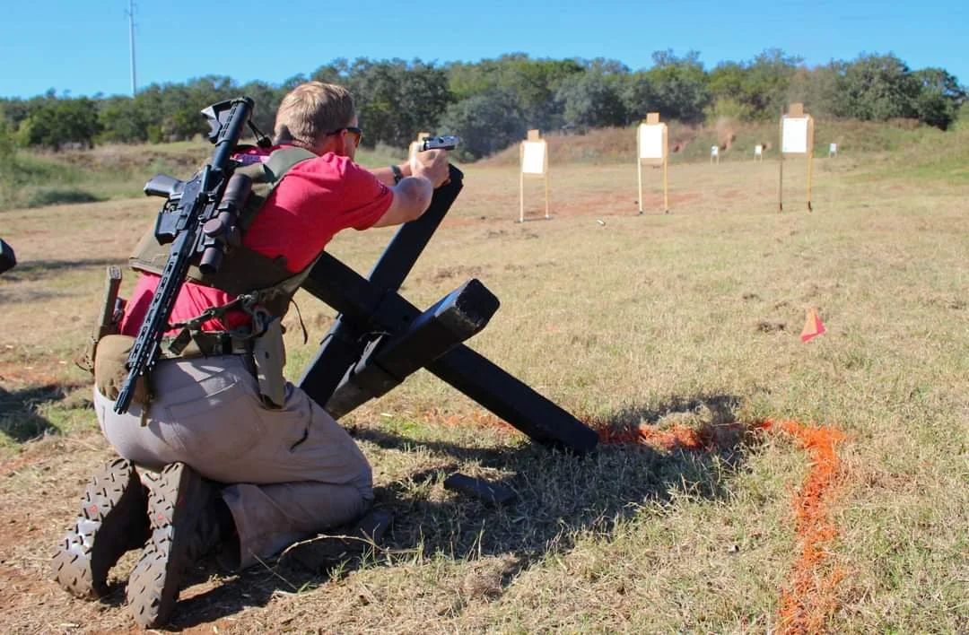 A man kneeling on the ground at a shooting range, aiming a pistol towards paper targets set up in the distance on a grassy field under a clear blue sky.