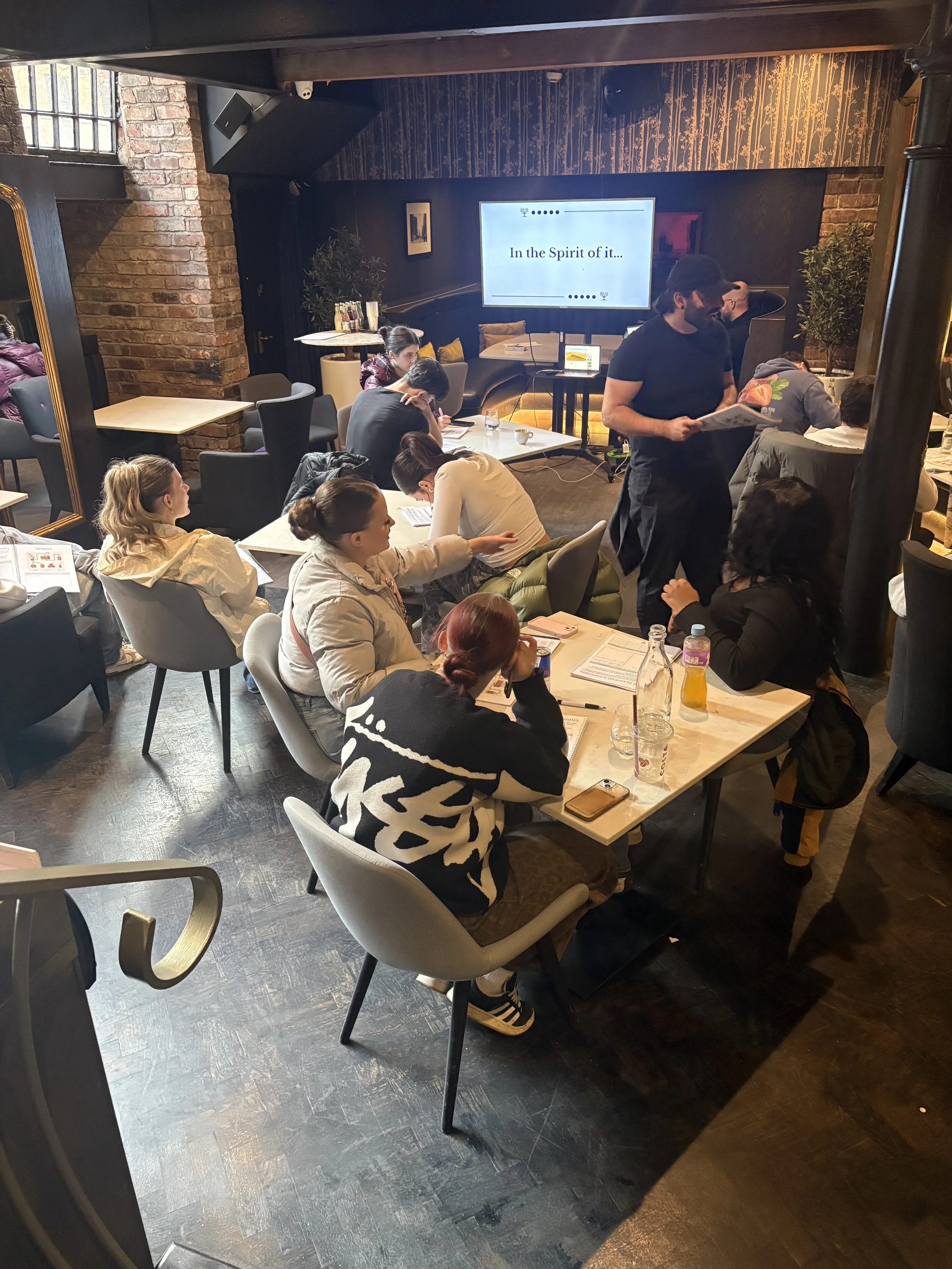 People attending a workshop or seminar in a cozy, dimly-lit coffee shop or cafe with brick walls and decorative wallpaper, sitting at tables with notebooks, drinks, and a large screen displaying the text "In the Spirit of it..."