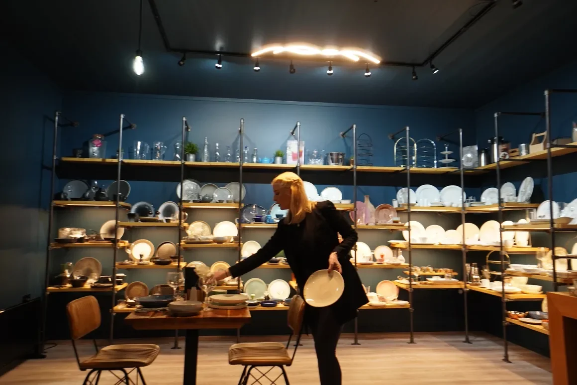 A woman in a black outfit organizing plates on a wooden table in a room with blue walls and shelves filled with dishes and glassware.