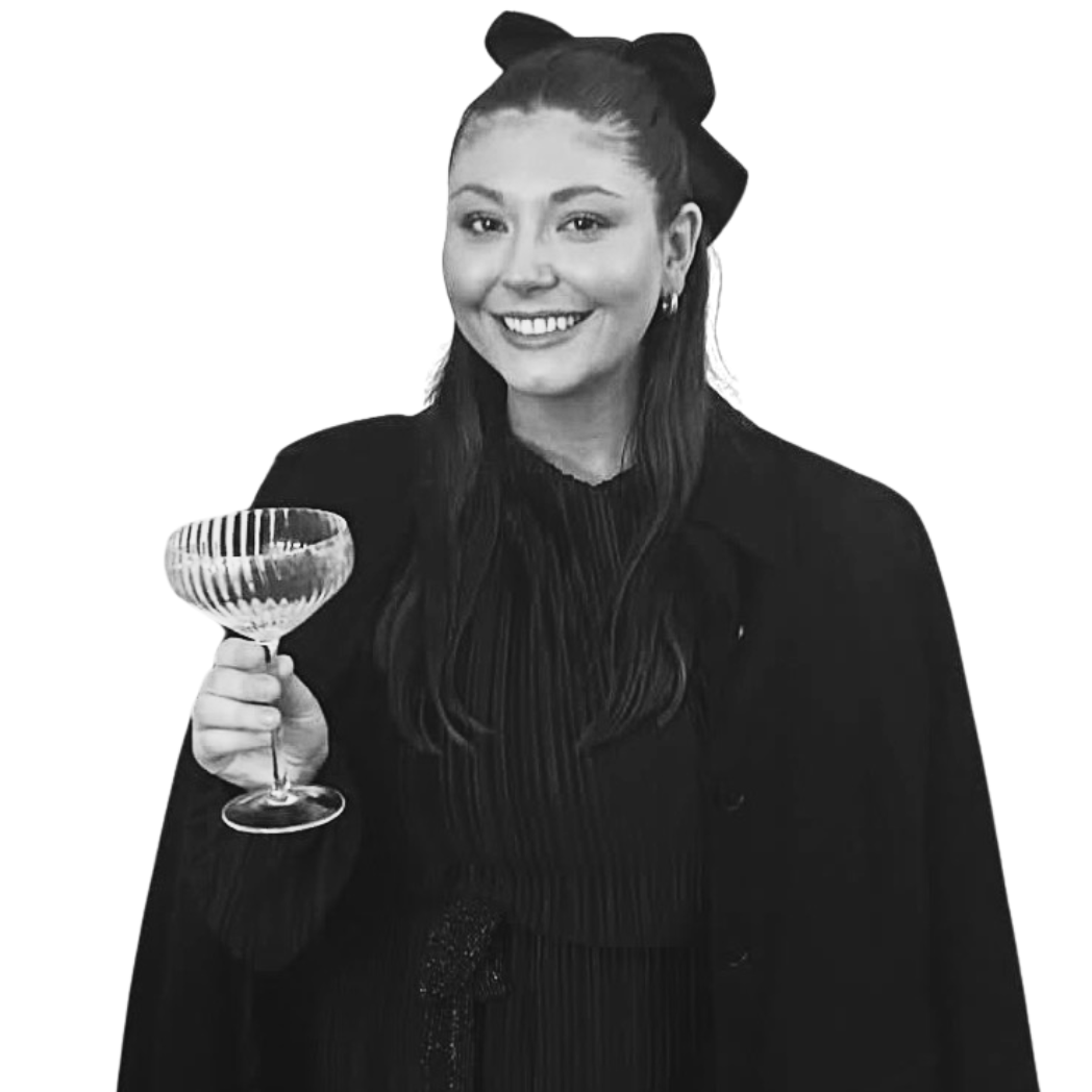 A young woman smiling and holding a glass of wine or champagne, wearing a black outfit with a bow in her hair.
