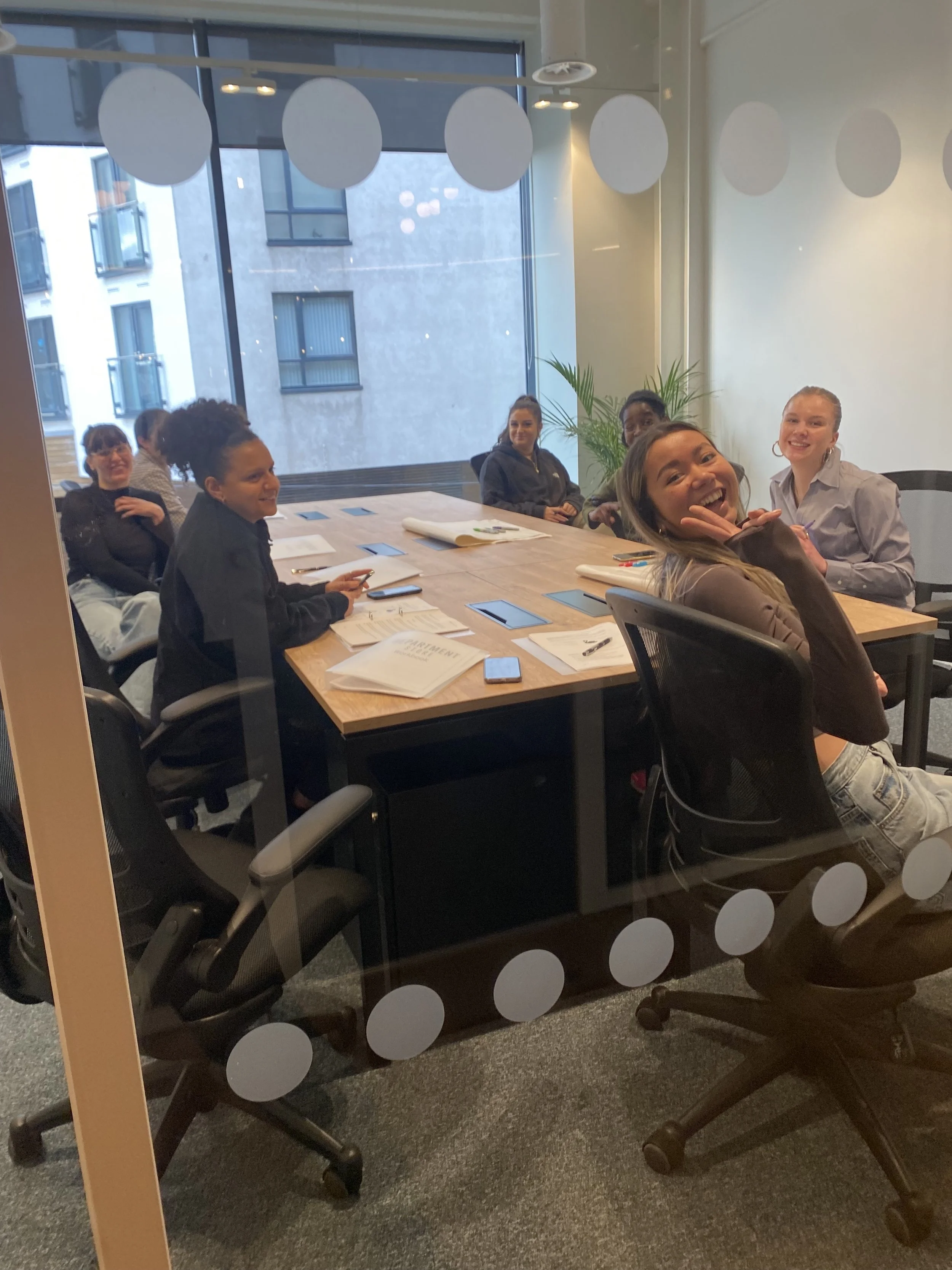A group of six women sitting around a conference table, smiling and looking through a glass door with white circular decals, in a modern office with large windows and a city view.
