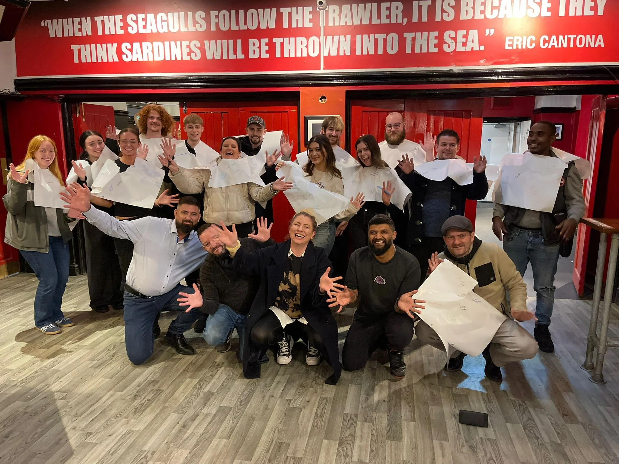Group of people smiling and posing playfully indoors, holding Fever Tree paper sheets with a quote from Eric Cantona displayed above them.
