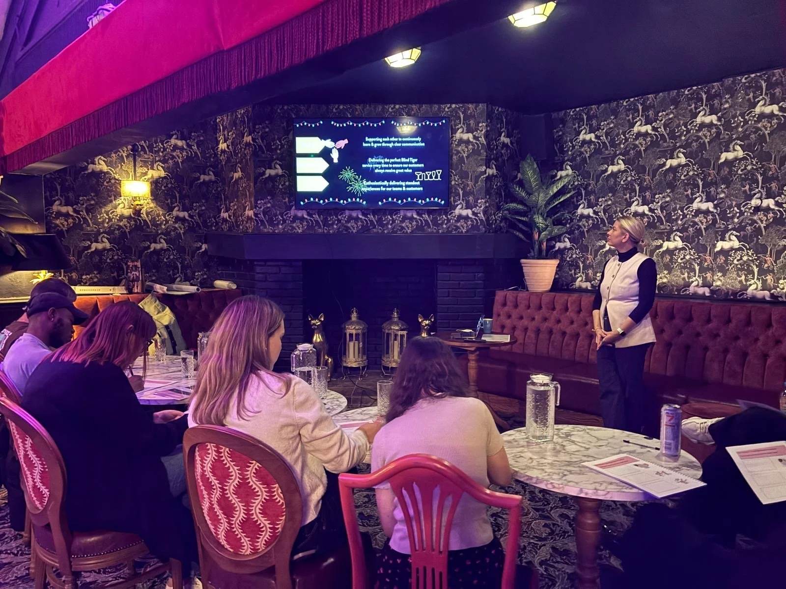 A woman stands on the right side of a room giving a presentation to a small audience seated at a round table with marble top. The room has dark patterned wallpaper, a large potted plant, and a tv screen displaying presentation slides. Water pitchers and documents are on the table.