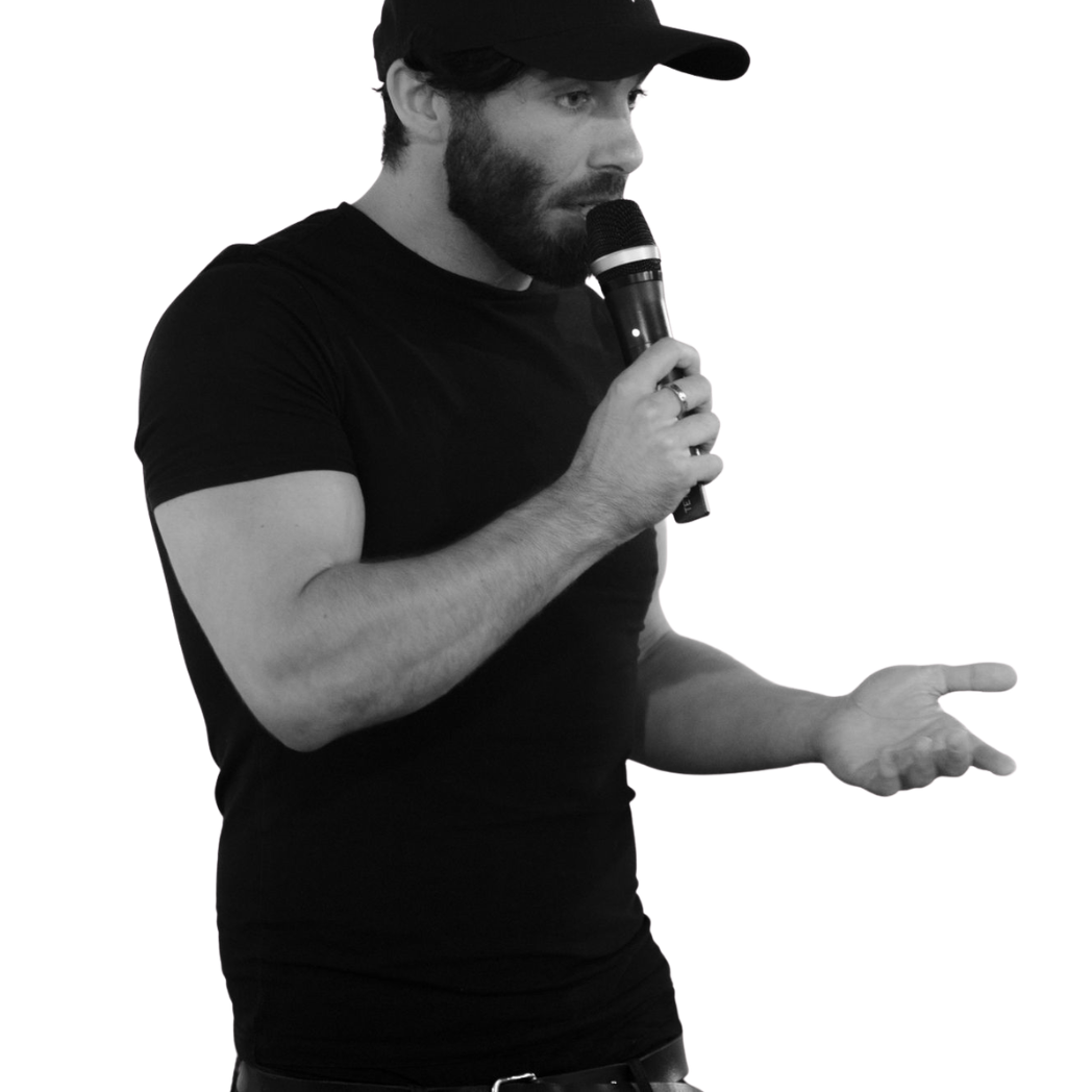 A man with a beard and wearing a black baseball cap and black t-shirt holding a microphone, gesturing with his other hand.