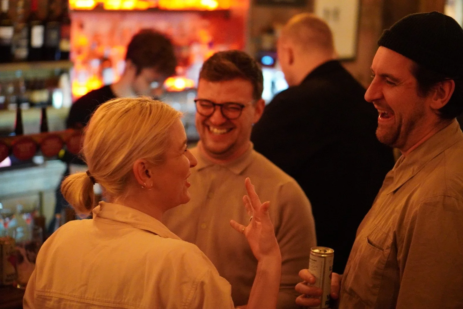 Four people laughing and talking in a dimly lit bar or pub, with shelves of bottles in the background.