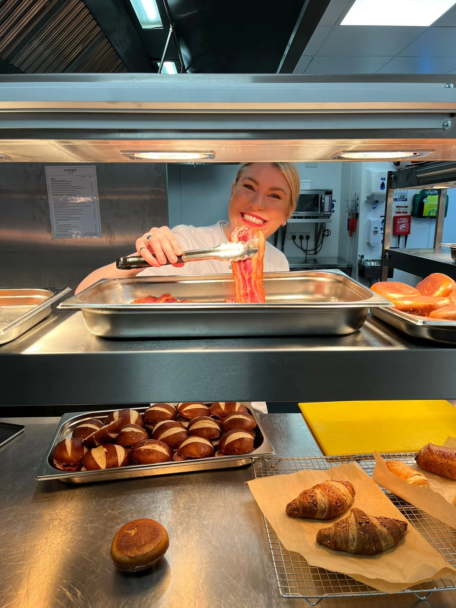 A smiling woman handling strips of bacon at a cafeteria serving counter, with baked goods like croissants and pastries visible on the counter below.