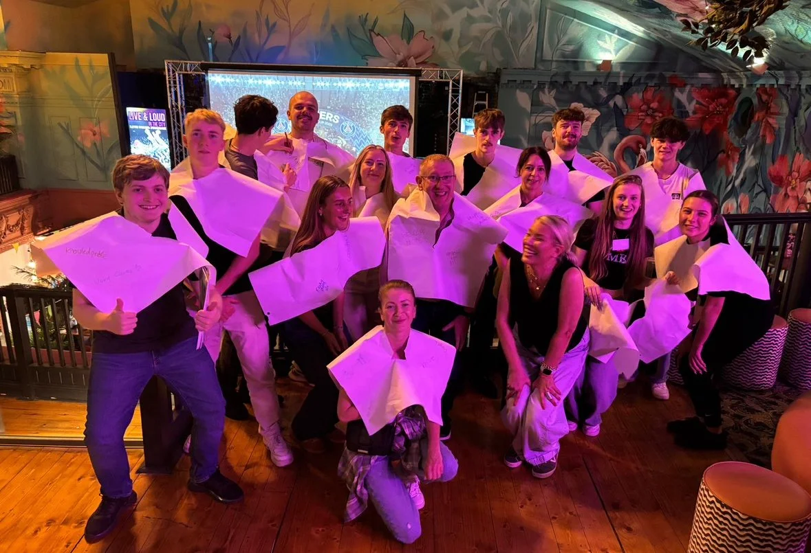 A group of people, mostly teenagers, wearing white napkins around their shoulders, are posing for a photo in a decorated indoor setting. Some are smiling and making gestures, with a man and a woman in the center. The background features a large floral mural and a screen displaying vibrant visuals.