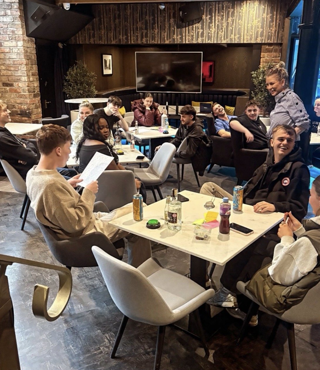 A group of young adults gather for New Opening Training, chatting and smiling in a relaxed, modern setting with drinks on the tables.