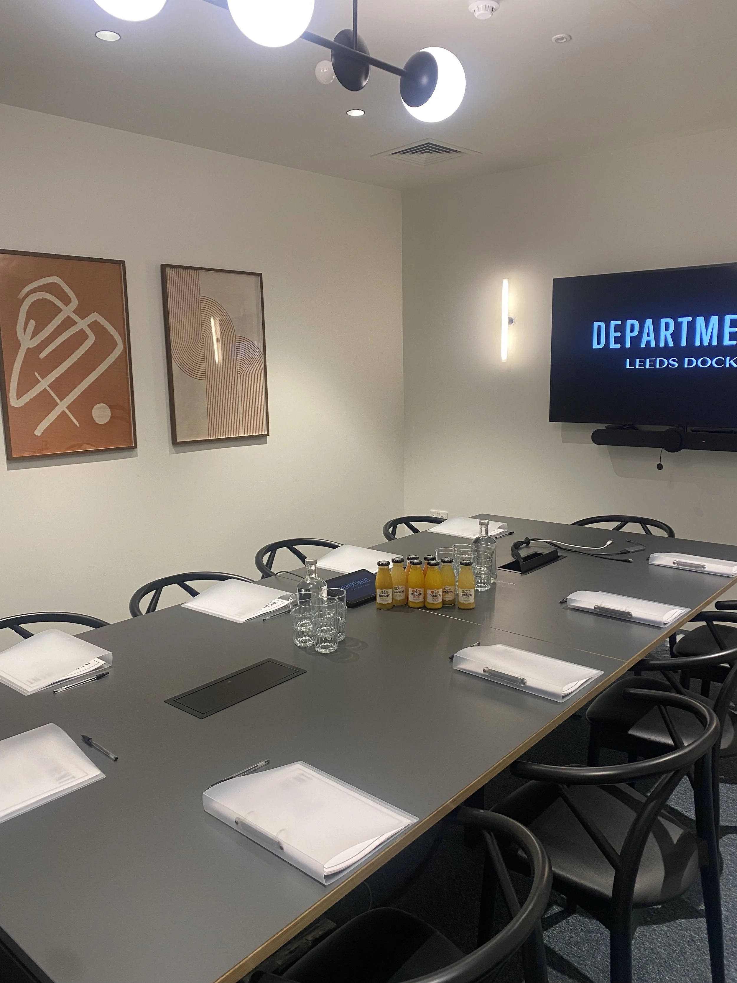 Meeting room with a long black table set with notepads, pens, glasses of water, bottles of juice, and electronic devices. On the wall, a mounted TV displays 'DEPARTMENT LEEDS DOCK'. The room has modern decor with abstract art on the walls and a ceiling light fixture.
