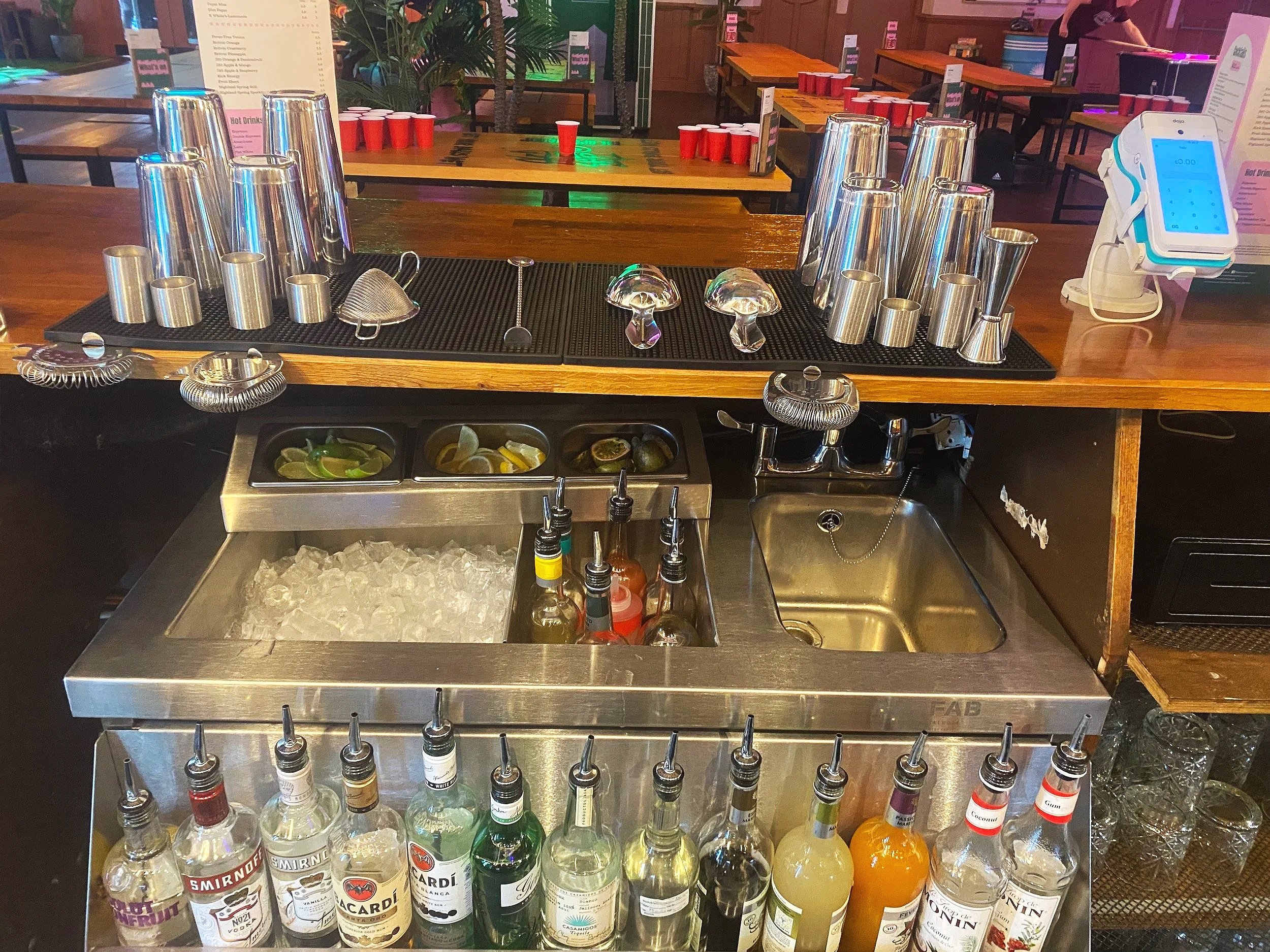 Bar counter with various liquor bottles, cocktail shakers, and drink-making tools, along with a sink and ice bucket, in a bar or restaurant setting.