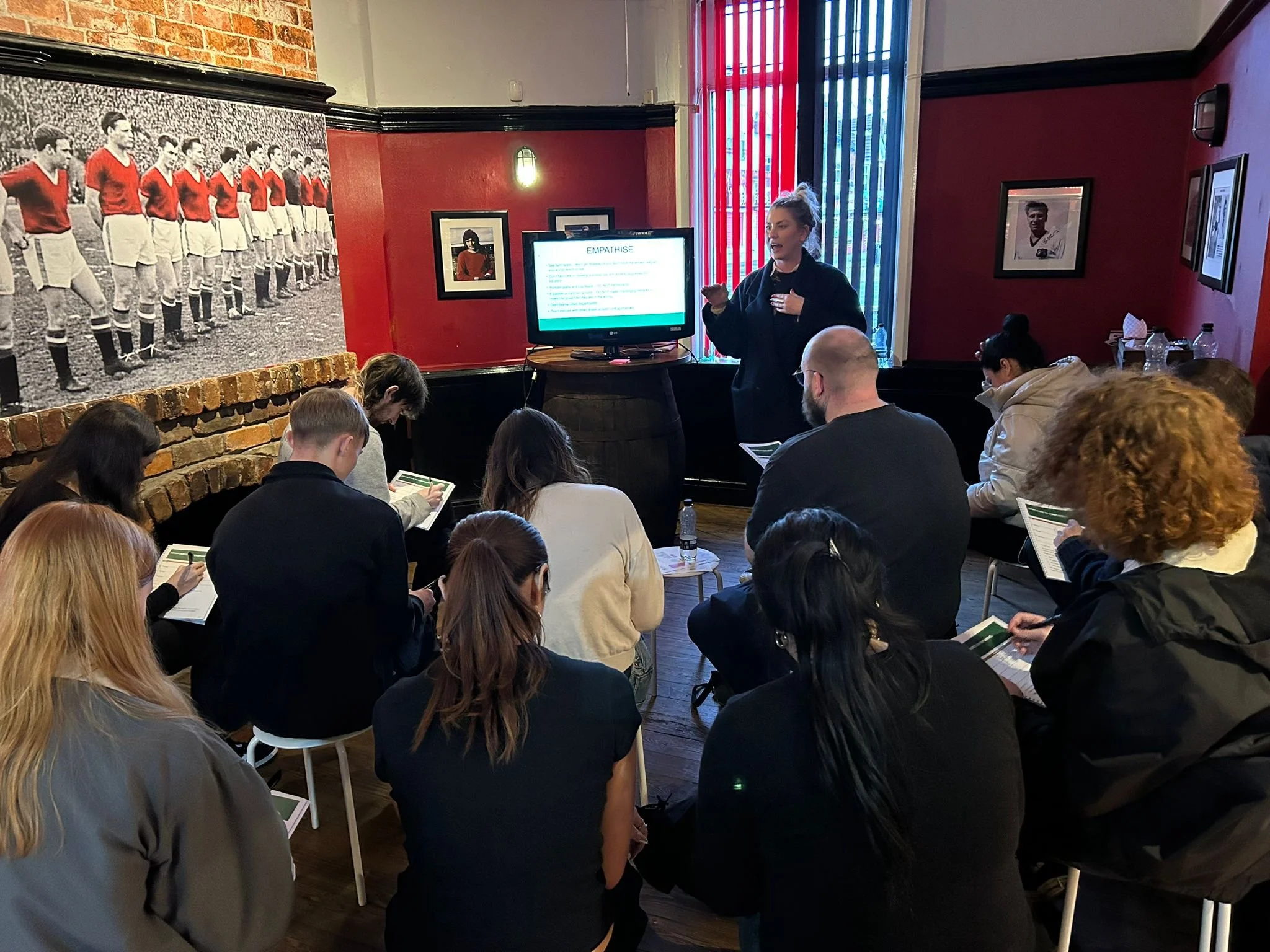 A group takes notes as a woman leads a Hospitality Training session in a room decorated with sports photos.