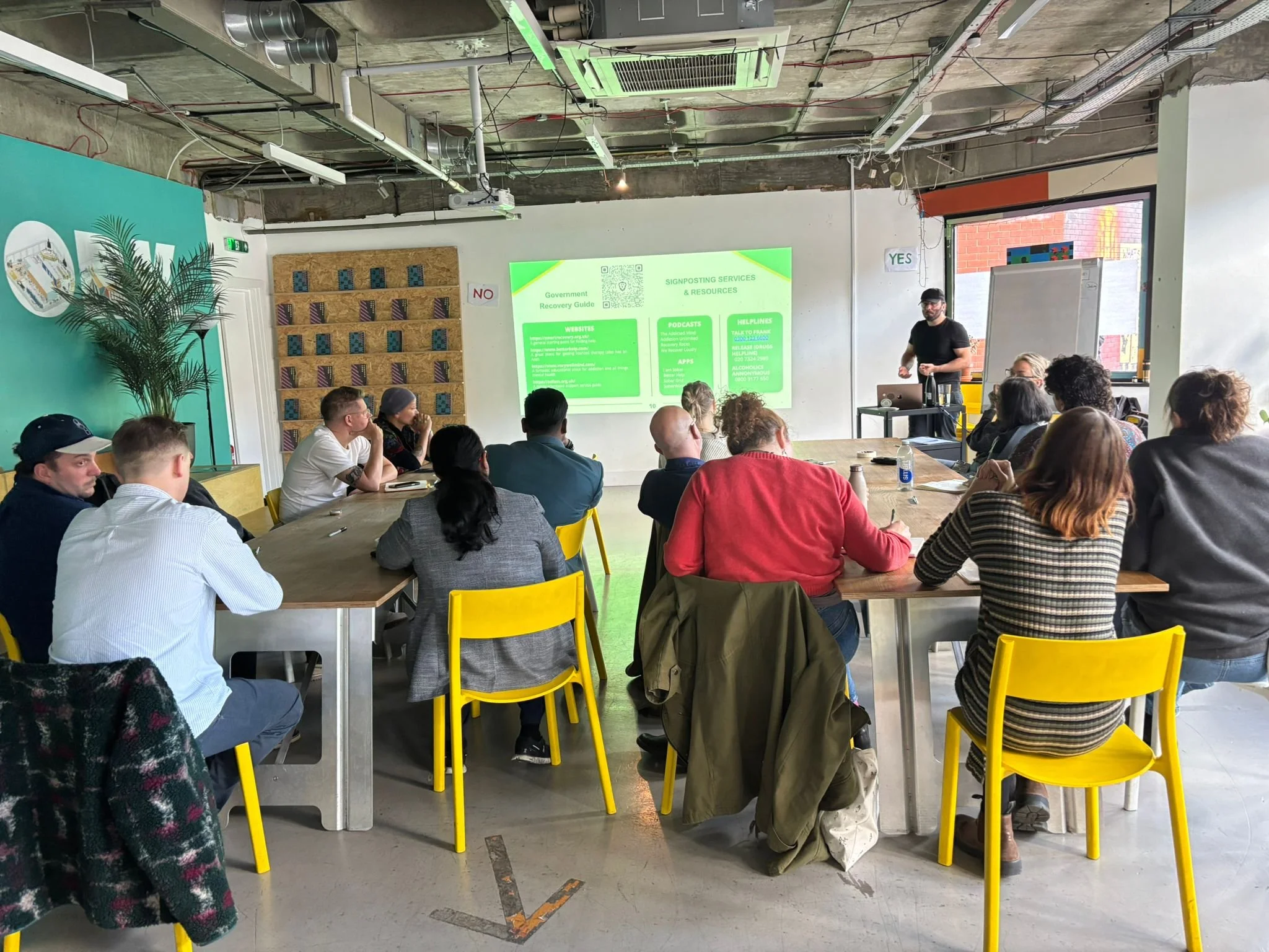 A group of people attending a presentation in a modern, industrial-style conference room with a large screen displaying information about government recovery guides and signposting services.