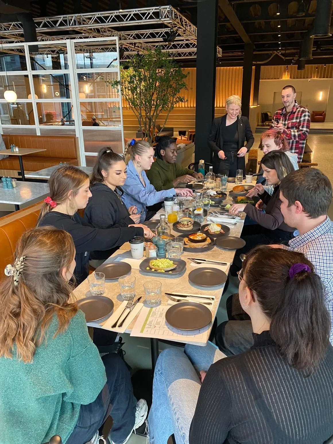 A group of people sitting at a long dining table in a modern restaurant, having a meal together. Two people are standing near the table, smiling and interacting with the group.