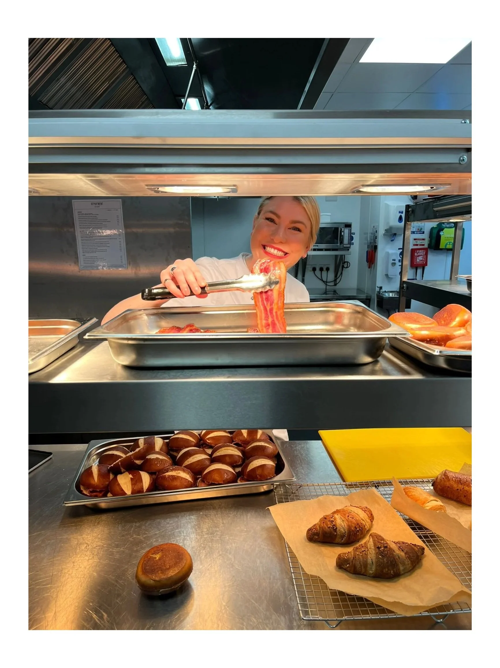 A smiling woman serving bacon from a hotel buffet station with baked goods including croissants and bread rolls on a tray and cooling rack in the foreground.