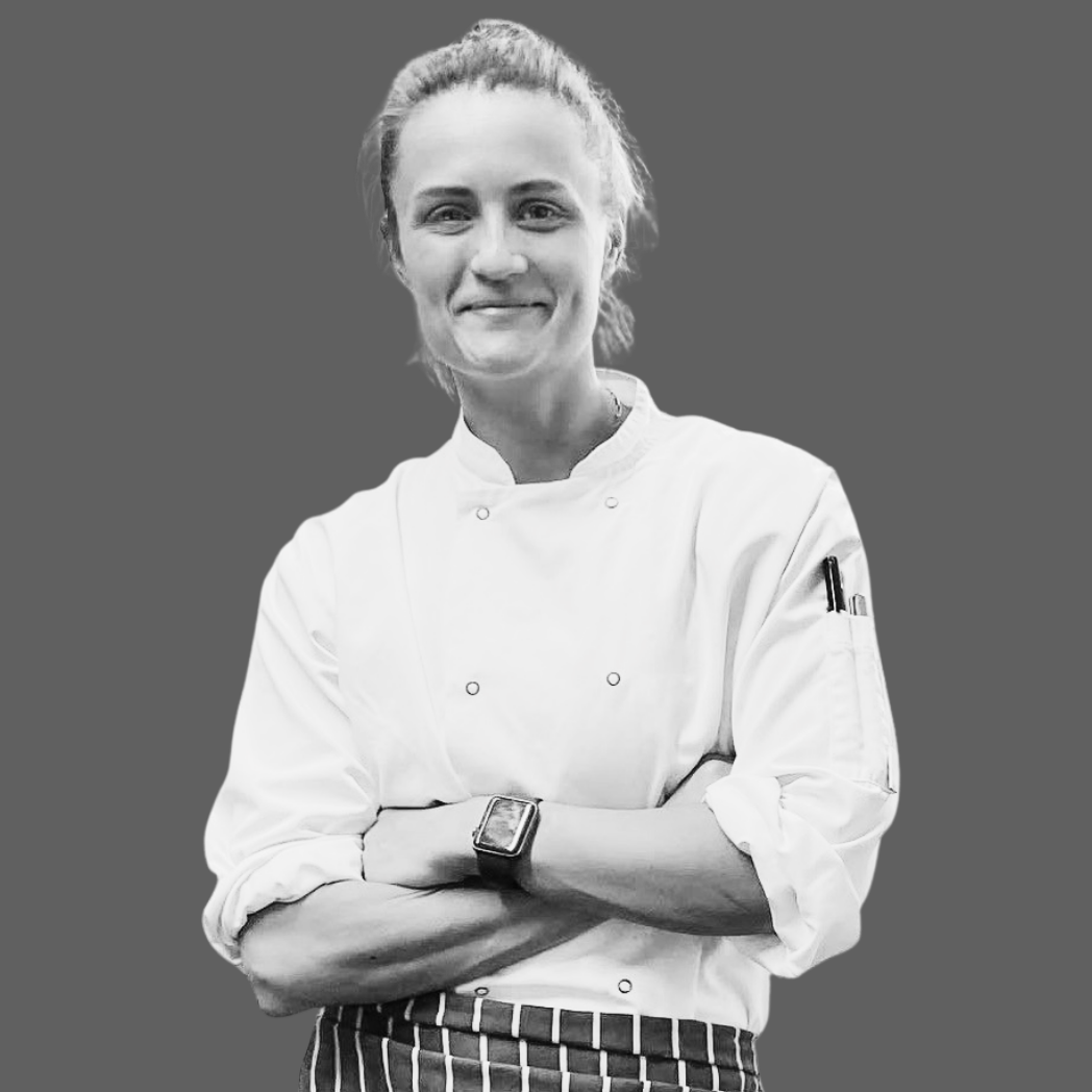 A female chef in a white chef's coat standing with her arms crossed, smiling, against a gray background.