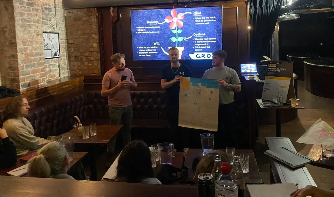 Three men presenting in a restaurant or bar setting, with a large screen behind them displaying a diagram of a flower with labeled sections. The audience is seated at tables with drinks, while one person is using a smartphone.