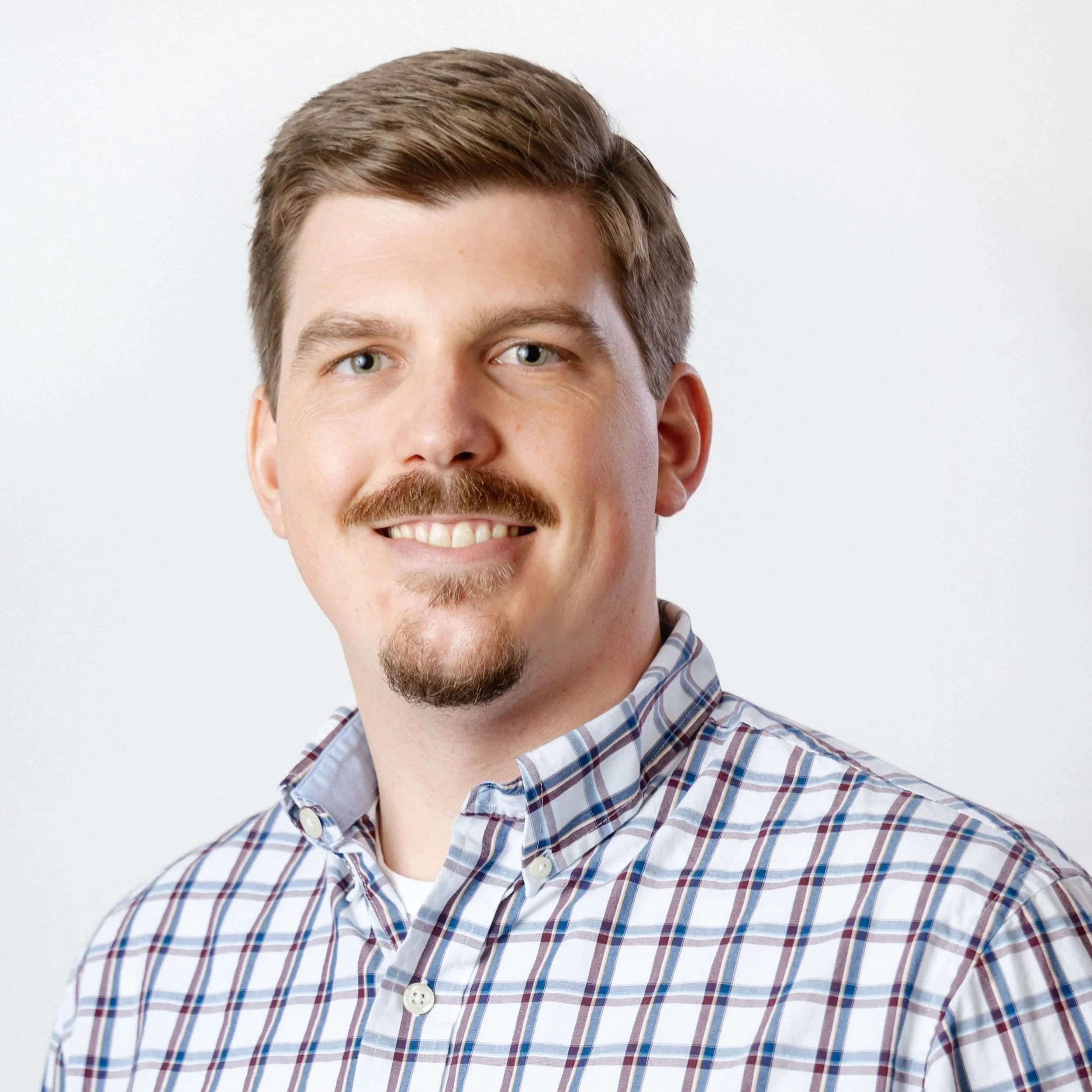 A smiling man with blue eyes, brown hair, a mustache, and a goatee, wearing a plaid button-up shirt.