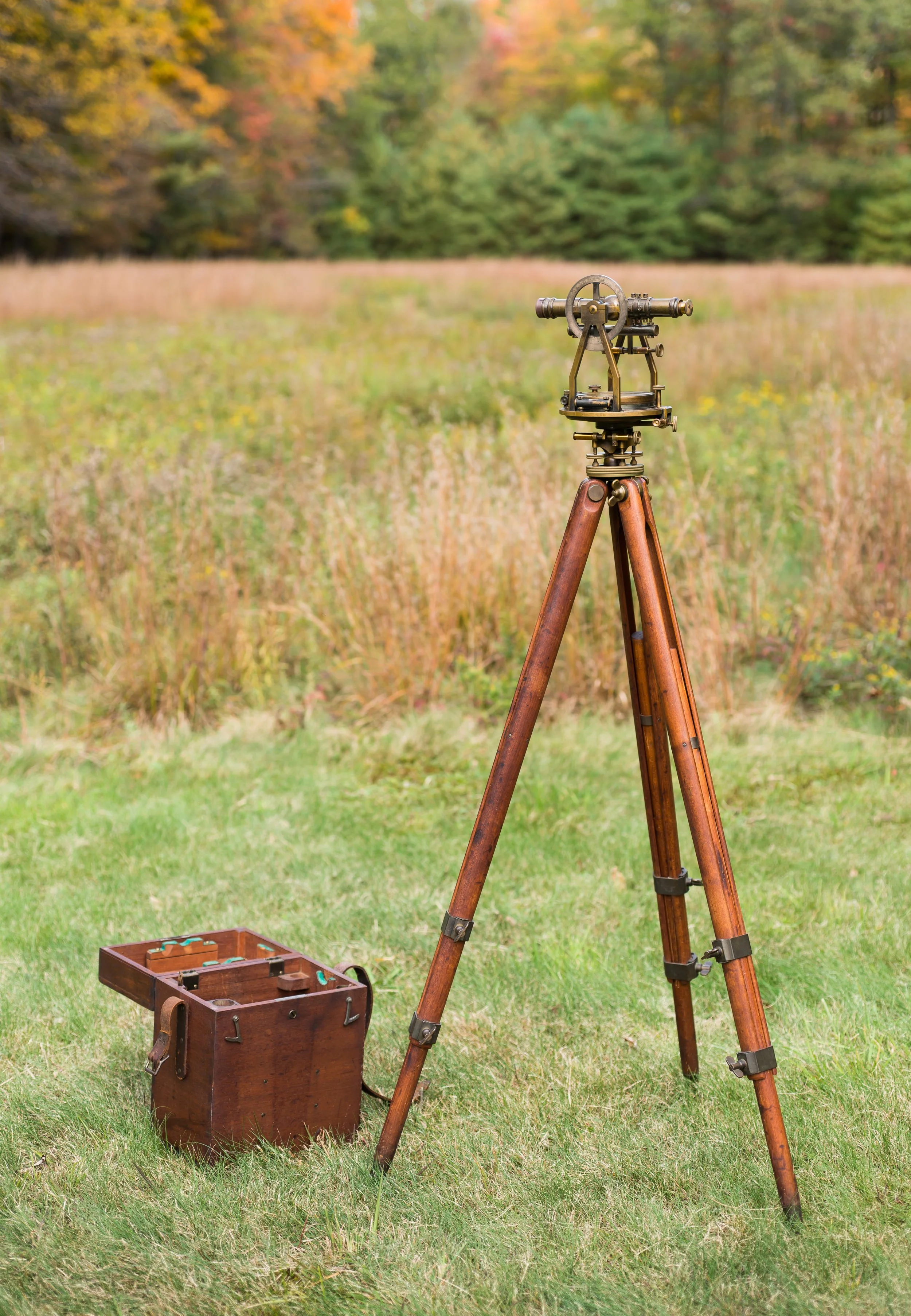 An antique brass theodolite mounted on a wooden tripod, set up outdoors on grass with trees and autumn foliage in the background.