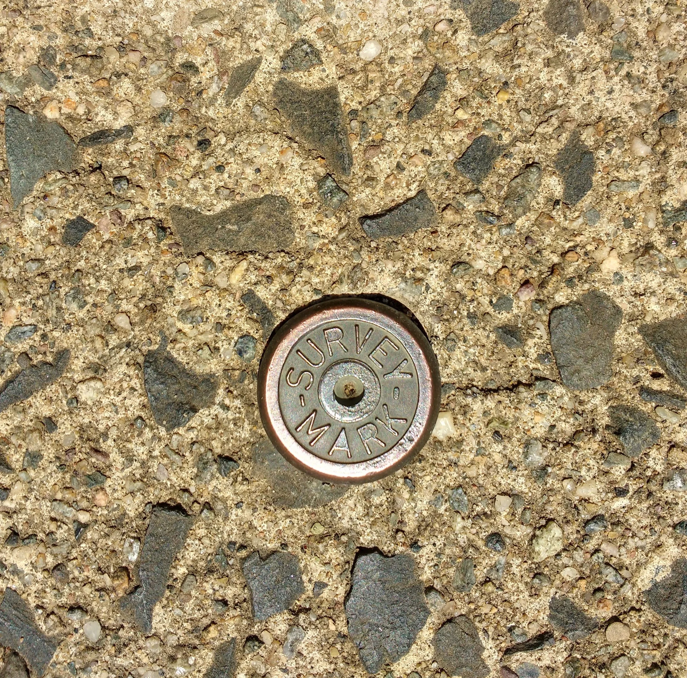 A metal survey marker embedded in a concrete sidewalk with small rocks.