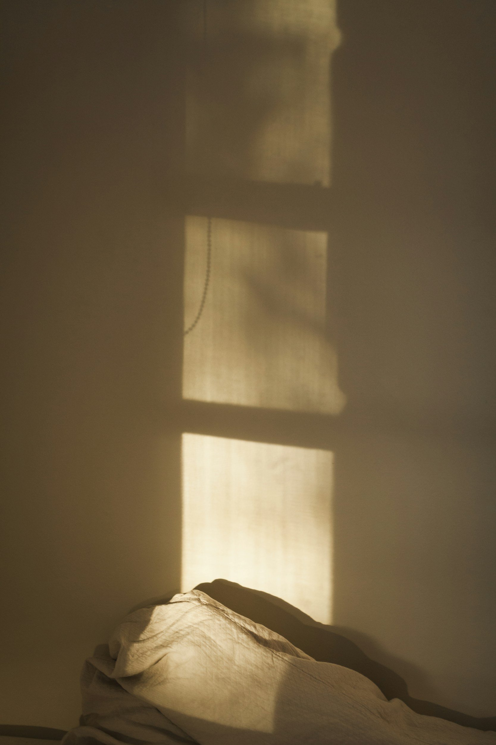 Sunlight casting rectangular shadow patterns on the wall from a window with blinds, with a rumpled bed in the foreground.