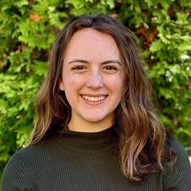 A woman with shoulder-length brown hair smiling outdoors in front of green foliage.