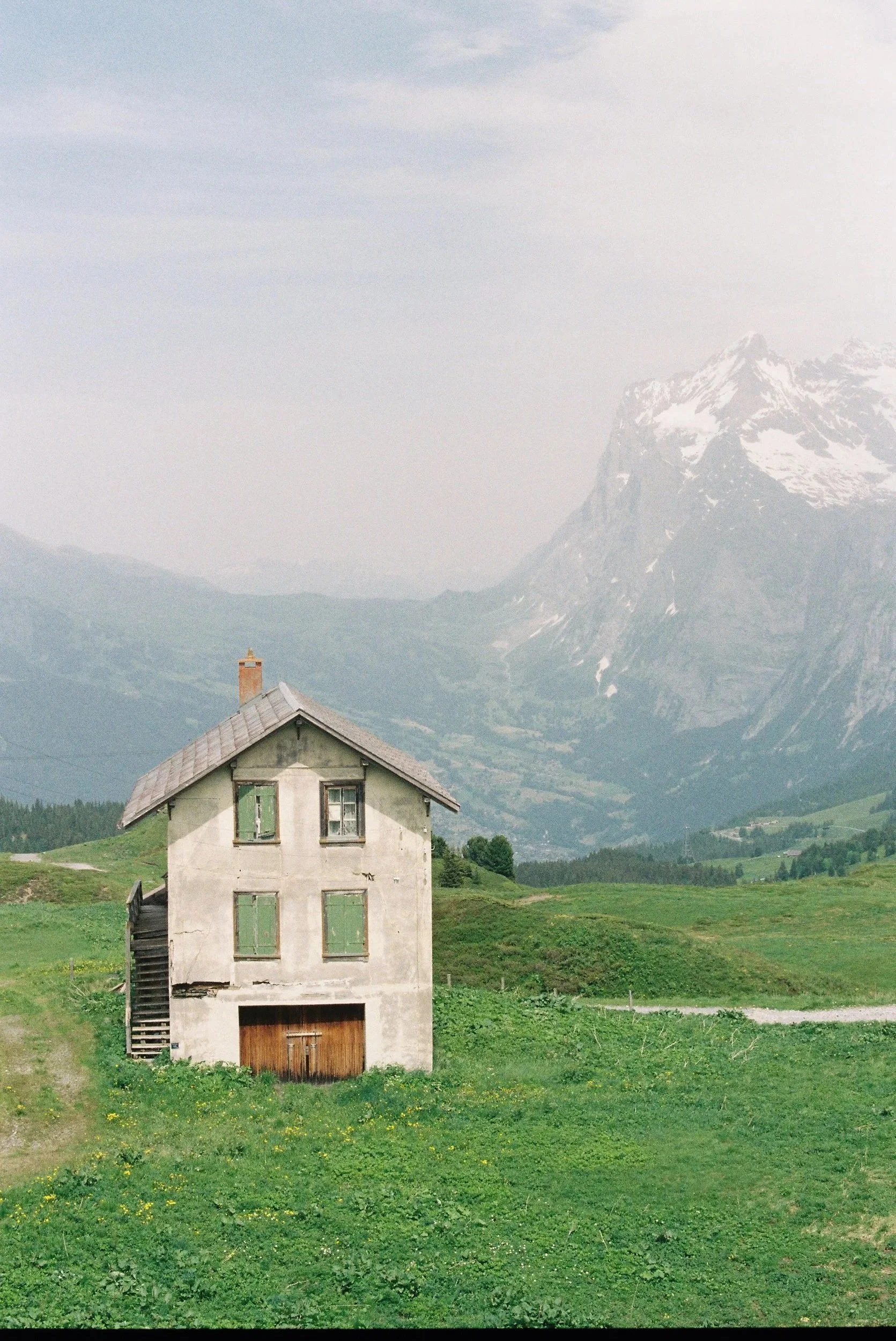 An old, weathered house with green shutters sits in a lush green field, with mountains in the background under a cloudy sky.