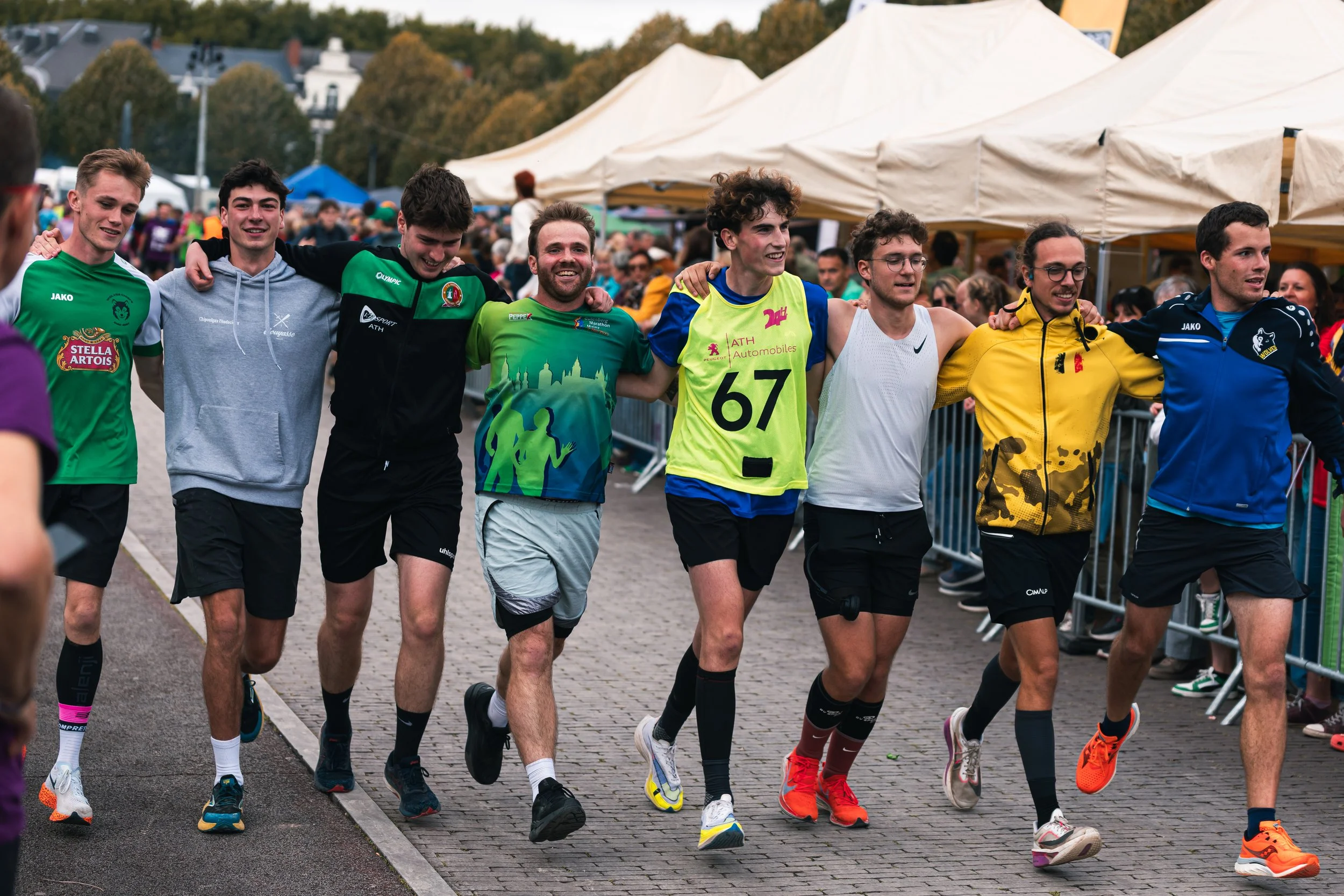 Un groupe de huit jeunes hommes, souriants, marchant ensemble lors d’un événement en plein air. Ils sont vêtus de vêtements de sport, certains portant des vestes ou t-shirts colorés, et ont des bras autour des épaules, montrant leur camaraderie. En arrière-plan, il y a des tentes et des spectateurs.