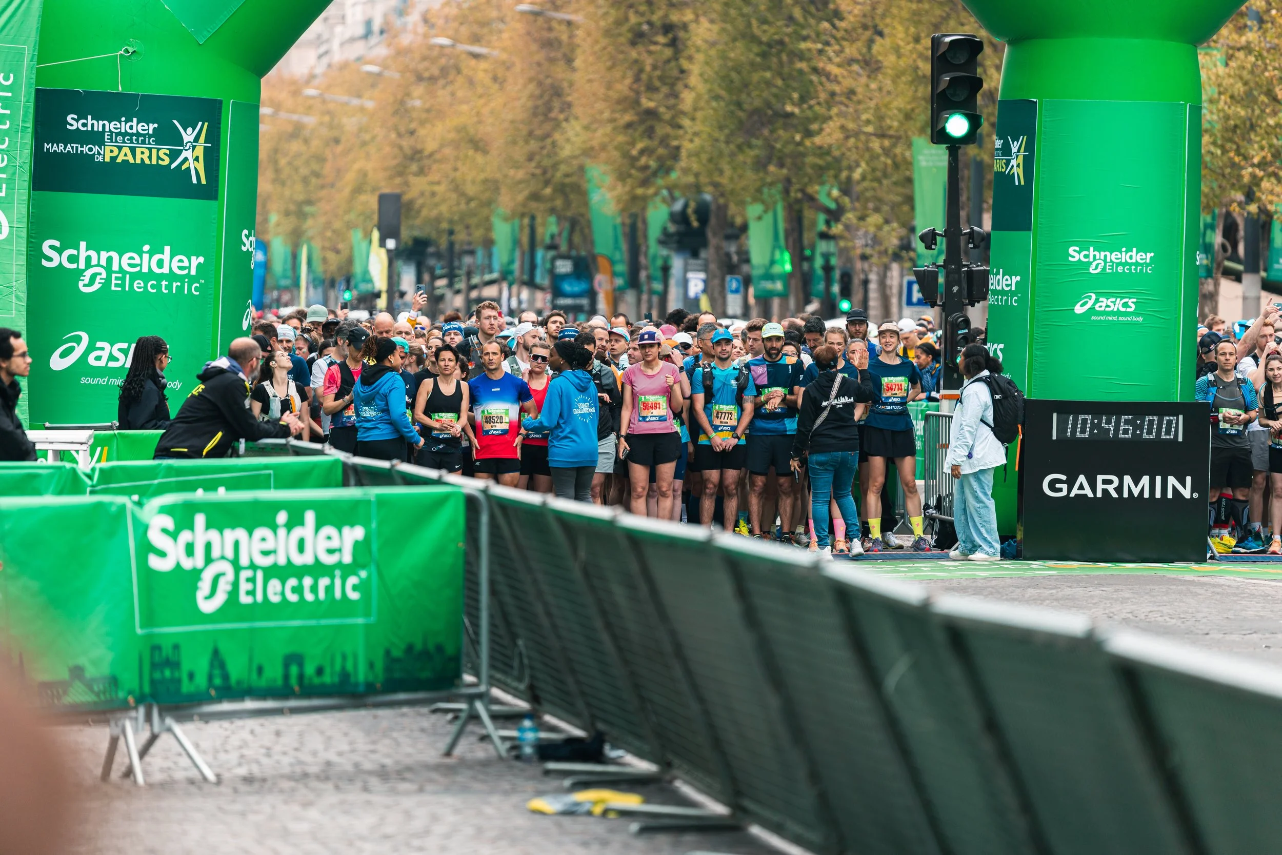 Départ de la marathon de Paris avec de nombreux coureurs derrière la ligne de départ. Infrastructure verte avec des sponsors comme Schneider Electric, Garmin, et Asics, et un écran digital affichant 10:46:00.