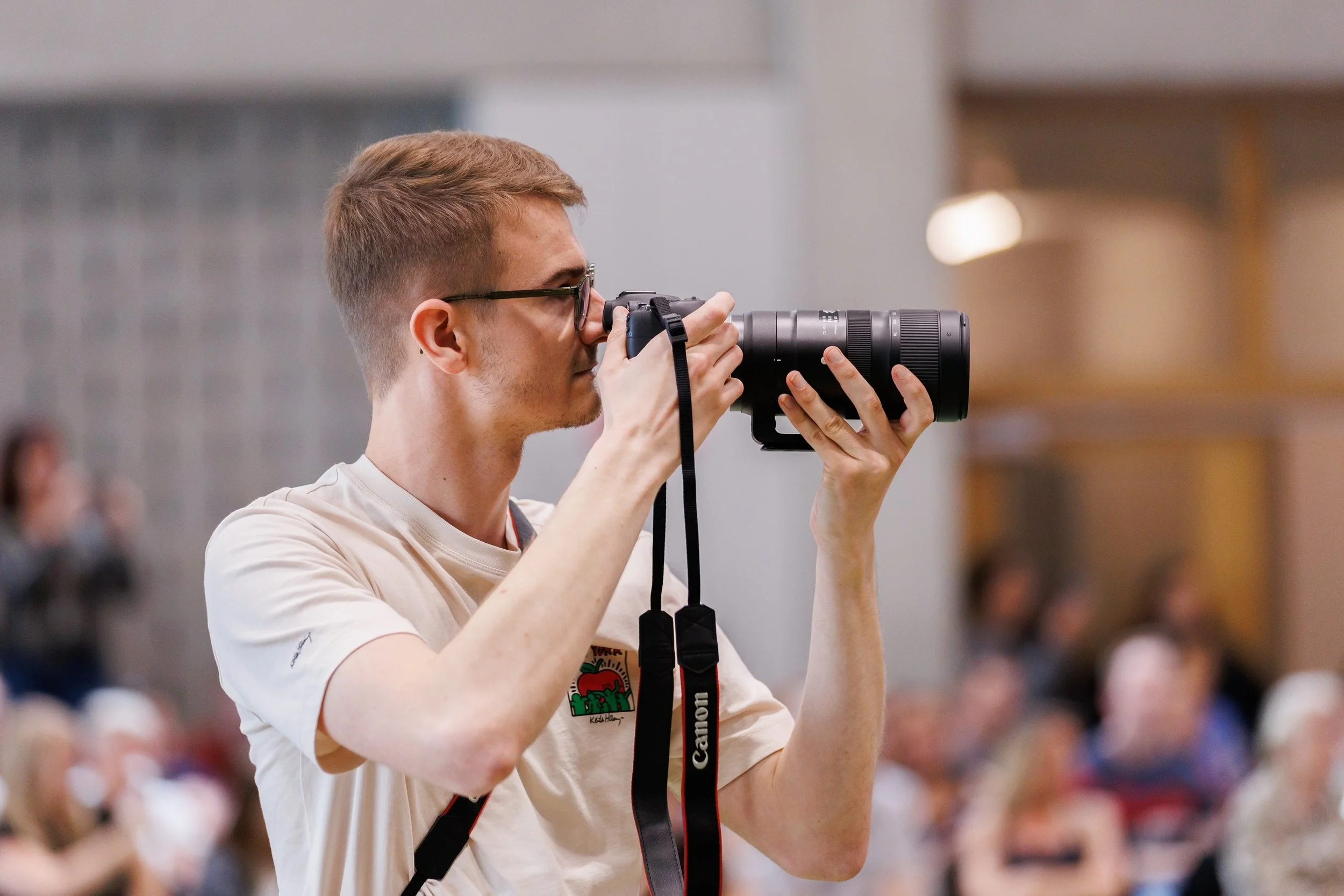 Jeune homme avec lunettes prenant une photo avec un appareil photo reflex dans un lieu public.