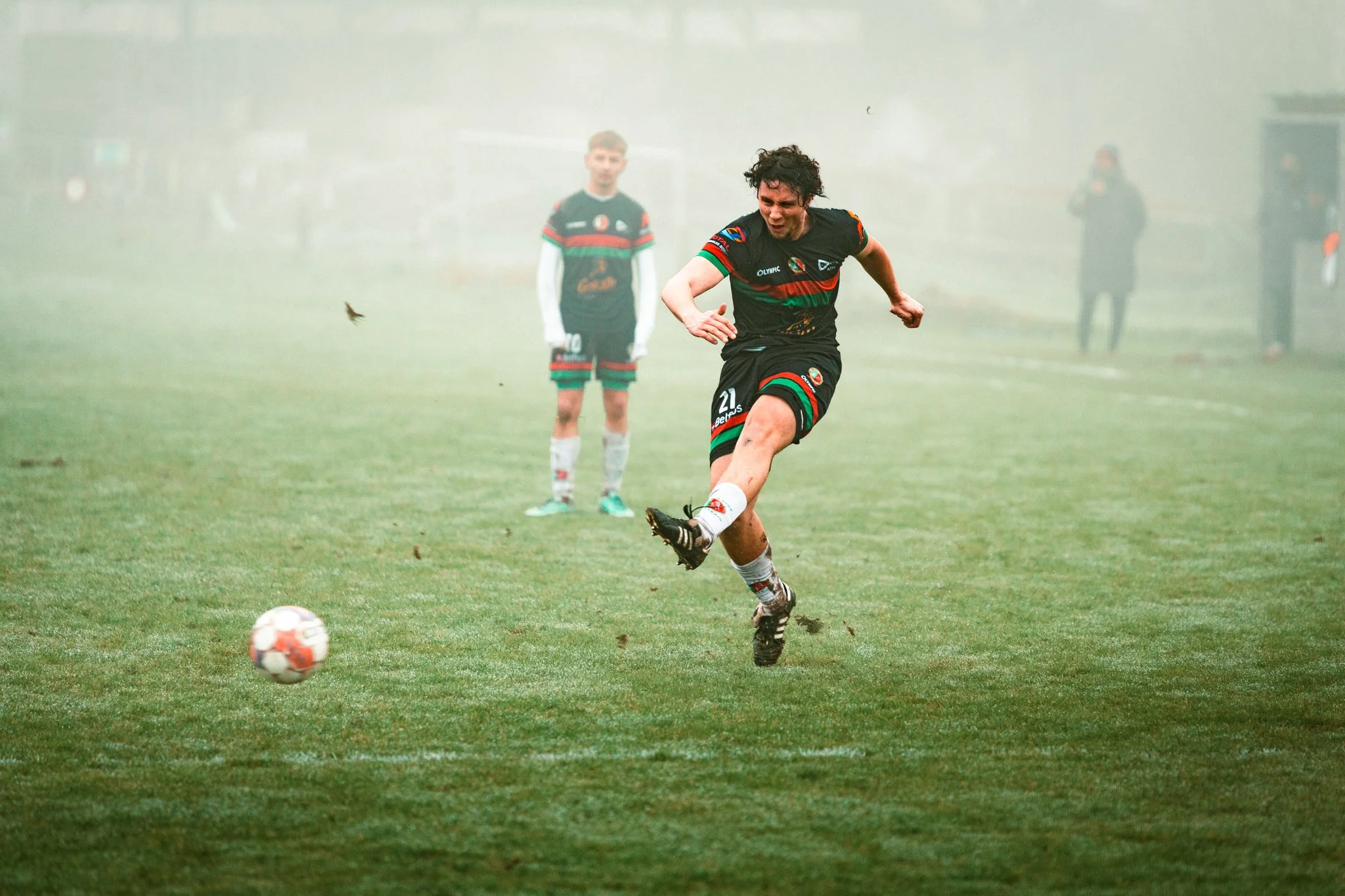 Un joueur de football en train de tirer un ballon lors d’un match sous une brume, avec deux autres joueurs en arrière-plan.