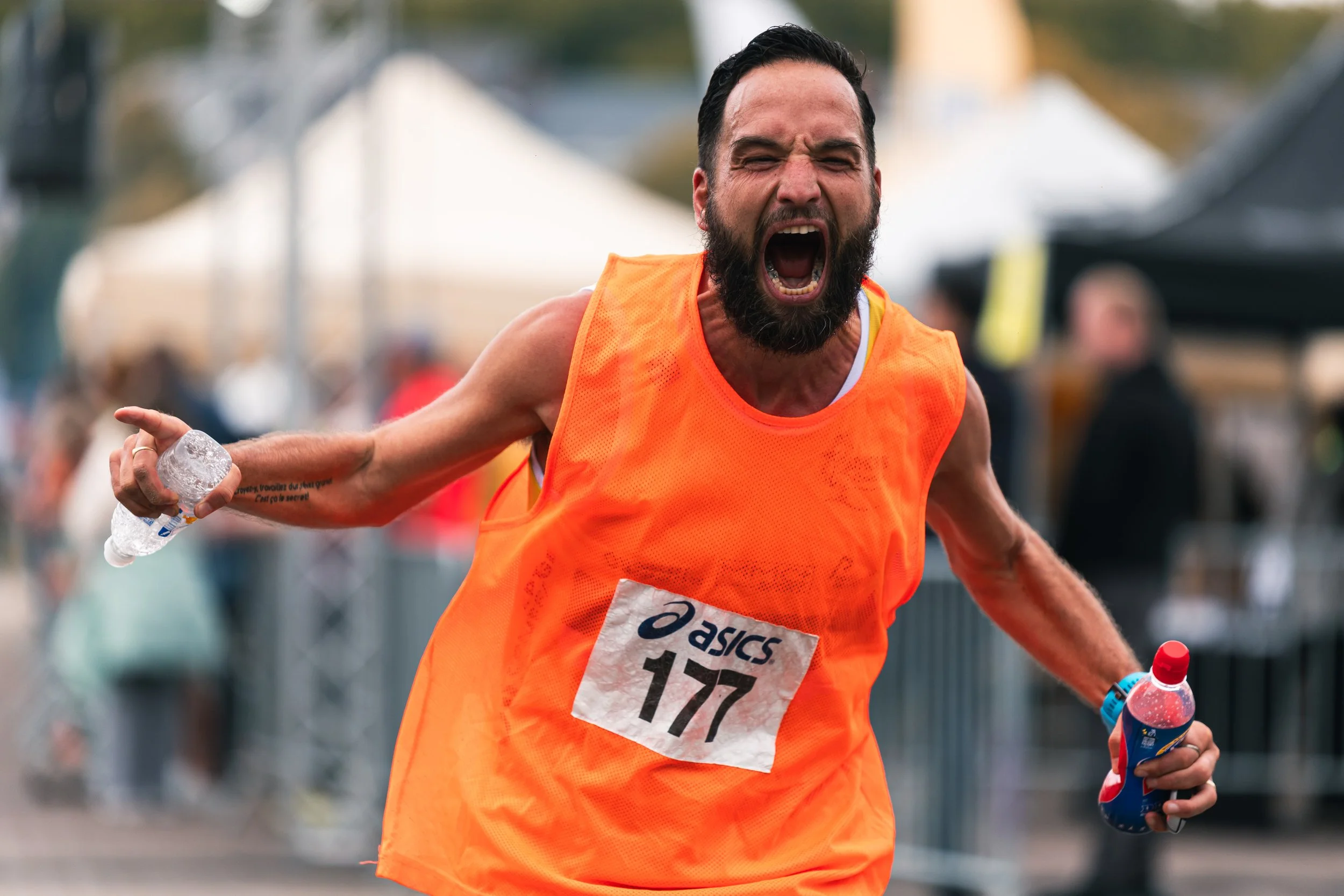 Un homme courant lors d'une course, crie de joie, porte un t-shirt orange avec un numéro 177, dans un environnement de marathon ou de course en extérieur.