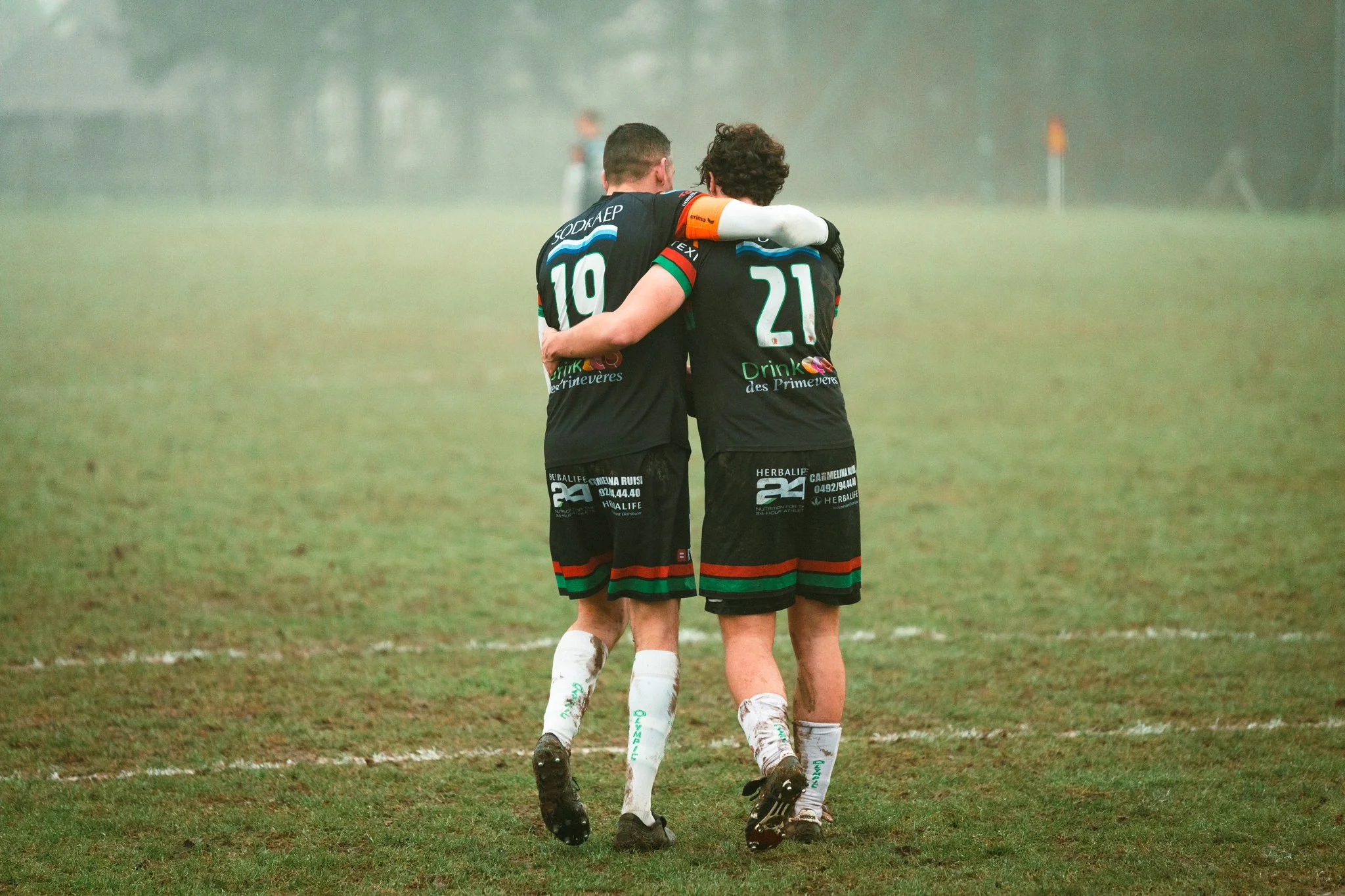 Deux joueurs de rugby en uniforme noir avec des numéros 10 et 21, s'enlaçant sur un terrain de rugby humide et brumeux.