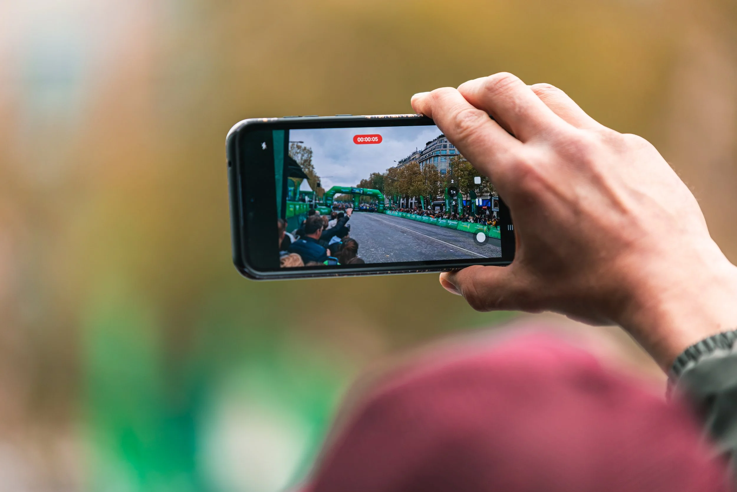 Une main tenant un smartphone filmant une course ou un événement sportif sur une rue urbaine, avec des spectateurs et des infrastructures en arrière-plan.